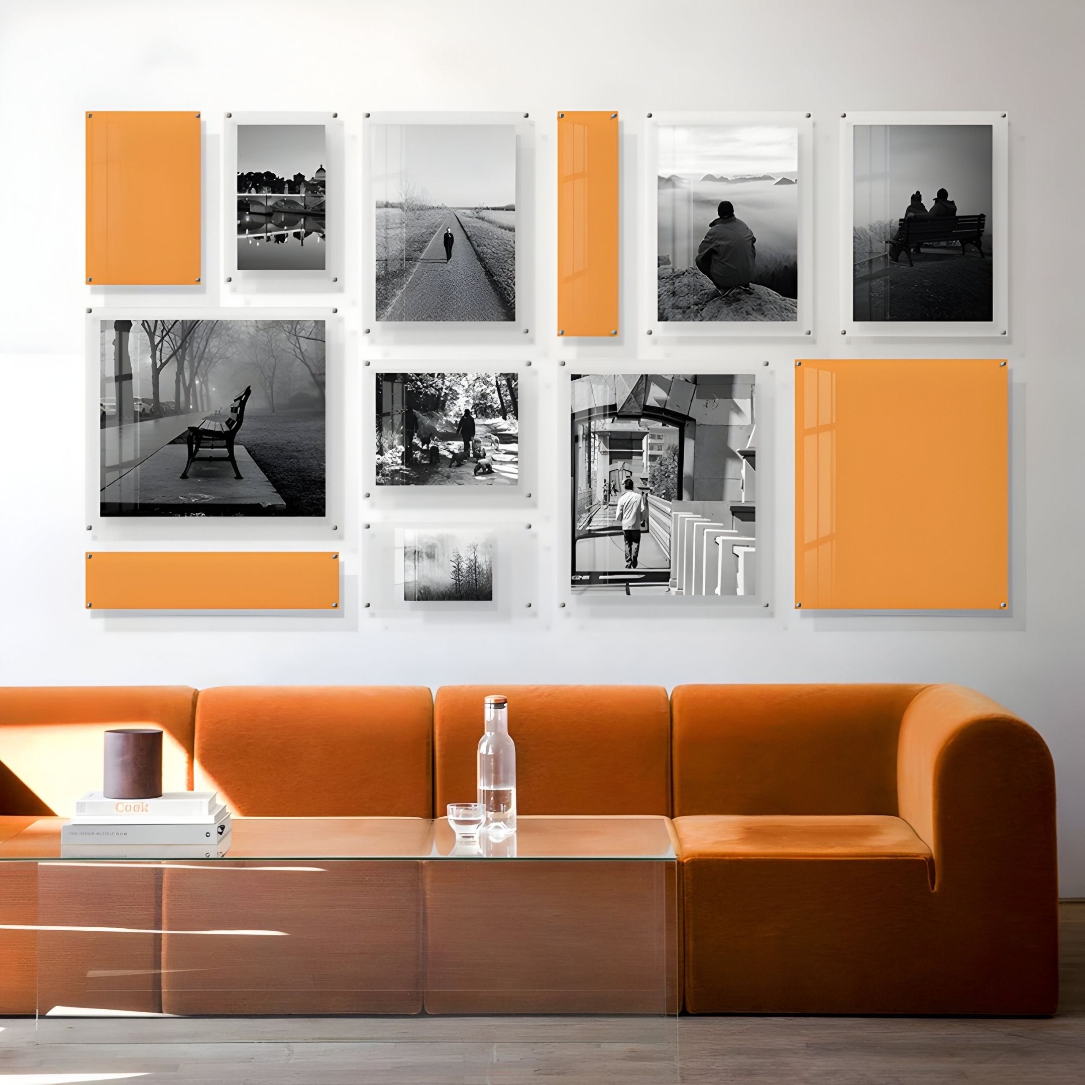 A modern living room features an orange sofa, glass coffee table, and the Giant Sculptures Framegrid Orange Mixed-Size Gallery 12-Set Acrylic Wall Art in a grid above the sofa. A glass water bottle and book on the table complete the contemporary decor.