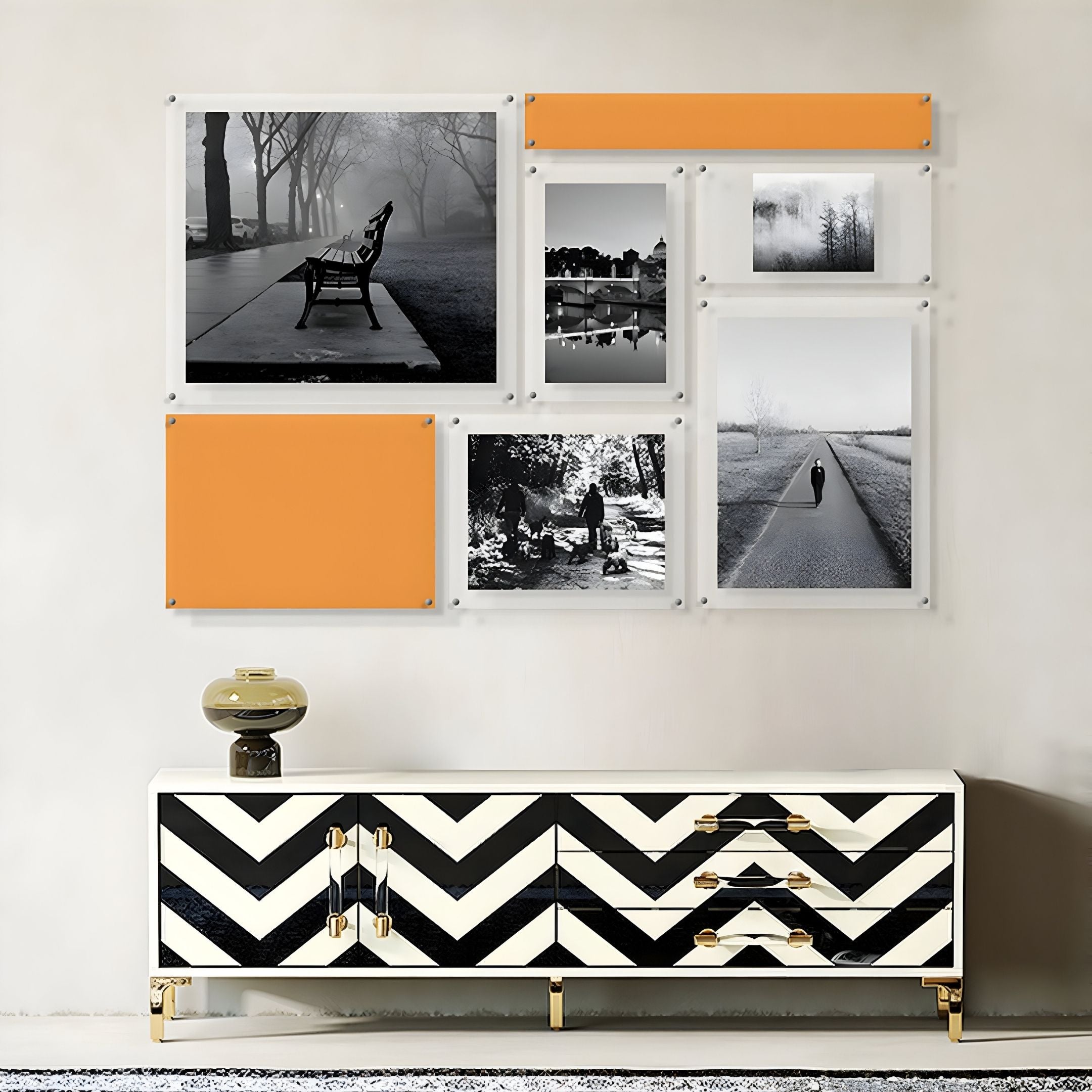 A modern living room features a chevron console table with gold accents, a round lamp, and Giant Sculptures’ Framegrid Orange Mixed-Size Gallery 7-Set Acrylic Wall Art displaying black-and-white photos and bold orange panels.