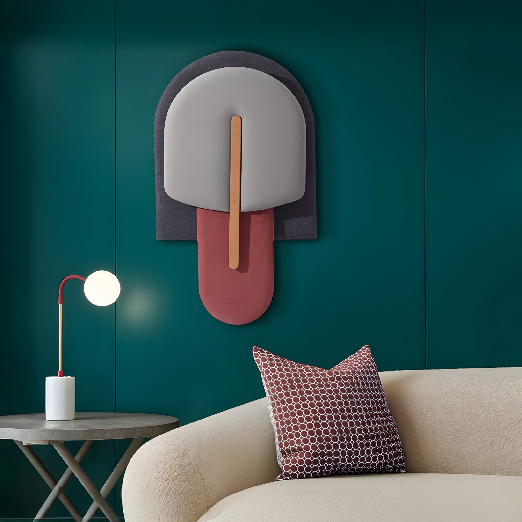 A modern living room with a teal wall, Giant Sculptures' Solvia Grey & Burgundy Layered Geometry 3D Wall Art, a round side table with a red and white lamp, and a cream sofa topped with a patterned brown pillow.