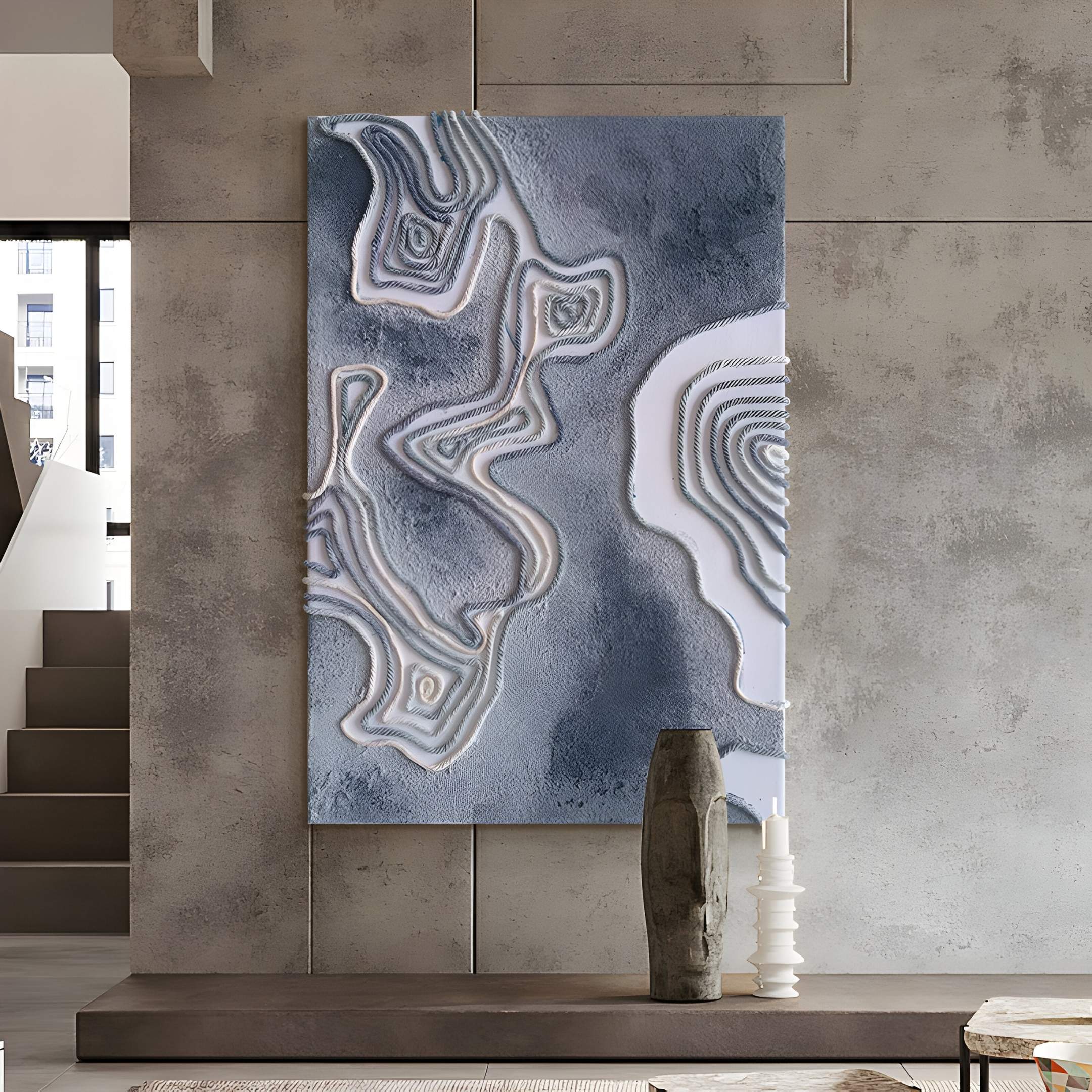 The Terracore Grey Contour Relief Abstract 3D Wall Art by Giant Sculptures hangs on a concrete wall, displaying hand-painted gray and white layers. Two sculptural vases below enhance the modern space with hints of stairs and windows in view.