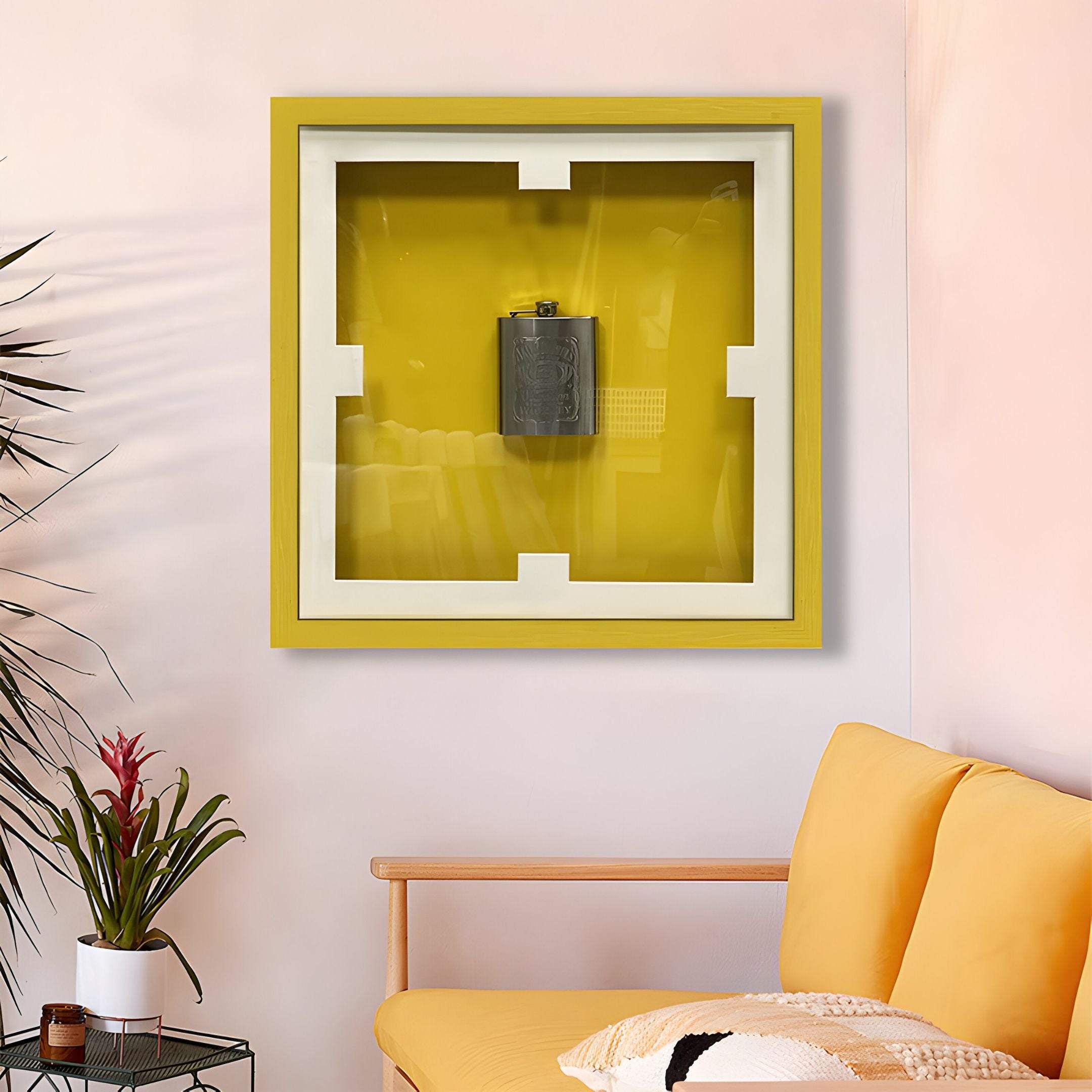 The Cavallero Yellow Hip Flask Display Framed Wall Art by Giant Sculptures hangs above a yellow couch on a light pink wall, with a potted plant on a side table, adding modern style to the room.