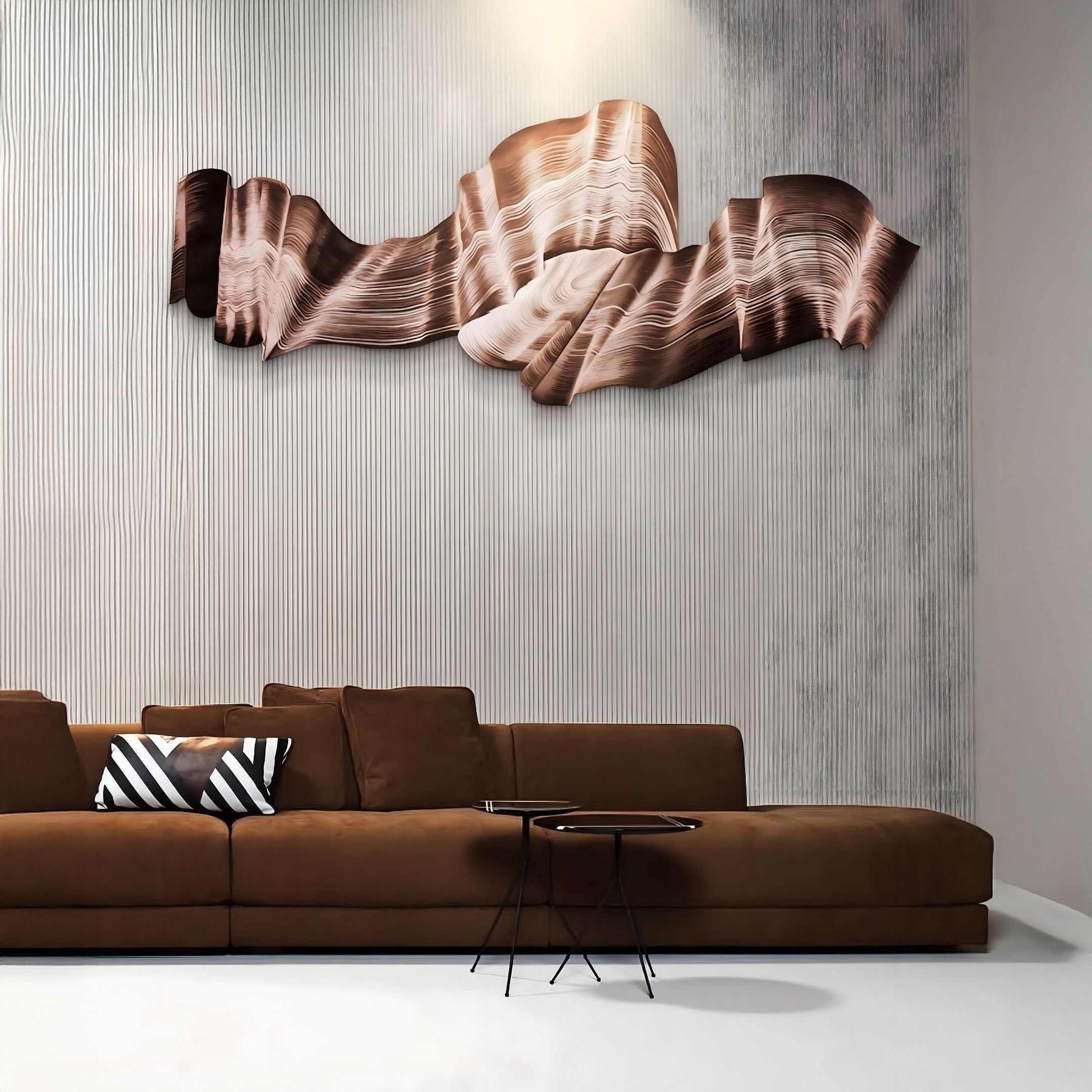 A modern living room with a brown sectional sofa, a black and white geometric pillow, two round nesting tables, and the Vellun Wave Bronze Drift Sculptural 3D Wall Art by Giant Sculptures as a striking feature against a textured gray wall.