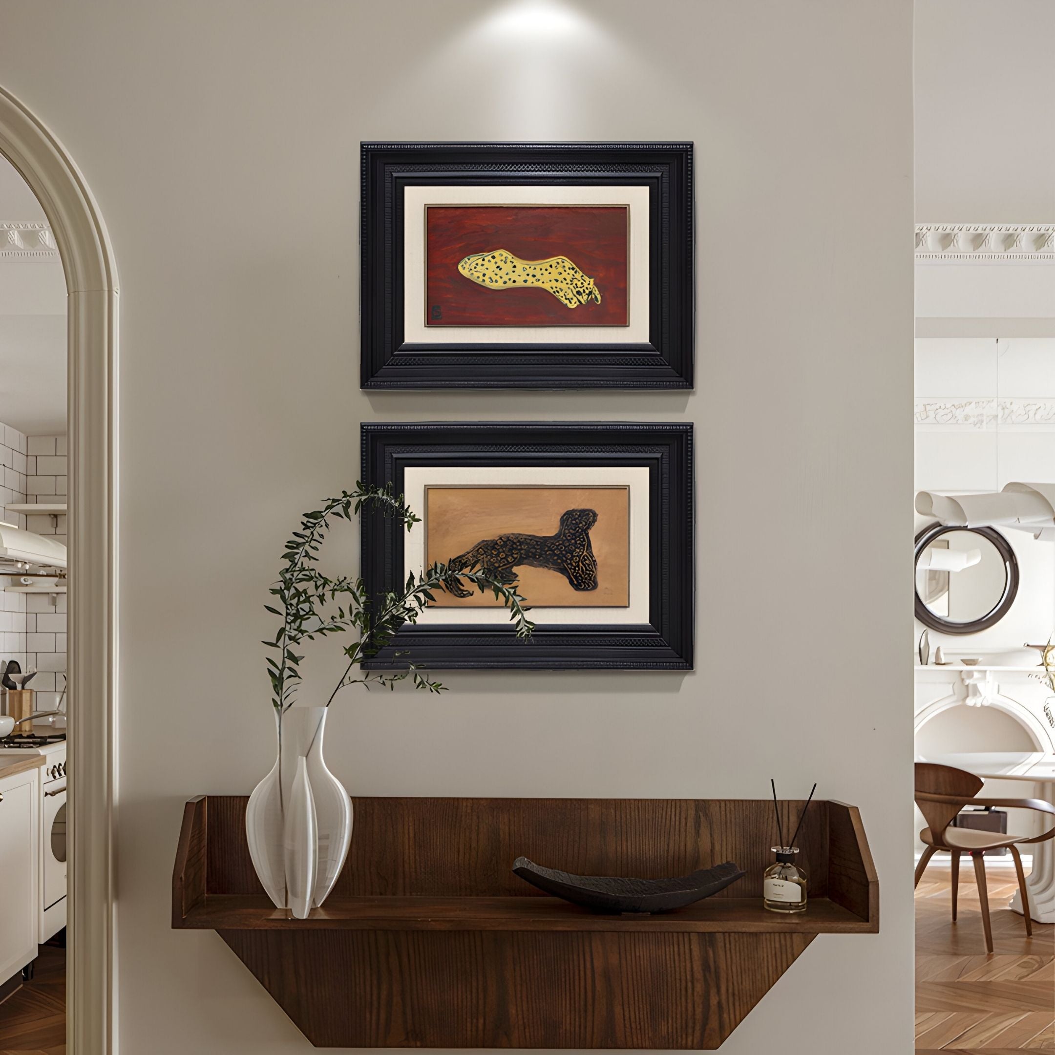 A modern hallway features the Kansora Leopard Emblem 2-Set Framed Wall Art by Giant Sculptures on a beige wall above a wooden floating shelf with a white vase of green branches and a small bowl; an arched doorway leads to the kitchen.