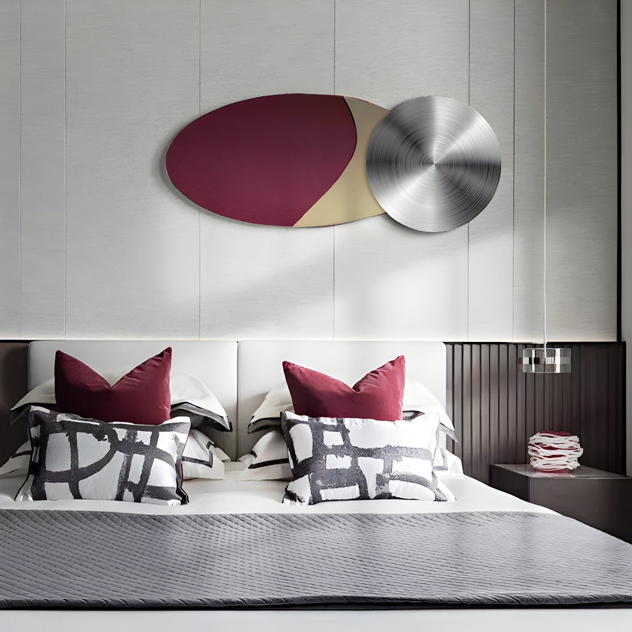 A modern bedroom with a large bed featuring black, white, and burgundy pillows, geometric bedding, and the Aurelune Burgundy & Taupe Silver Metal Disc 3D Wall Art by Giant Sculptures above the headboard for a stylish contemporary touch.