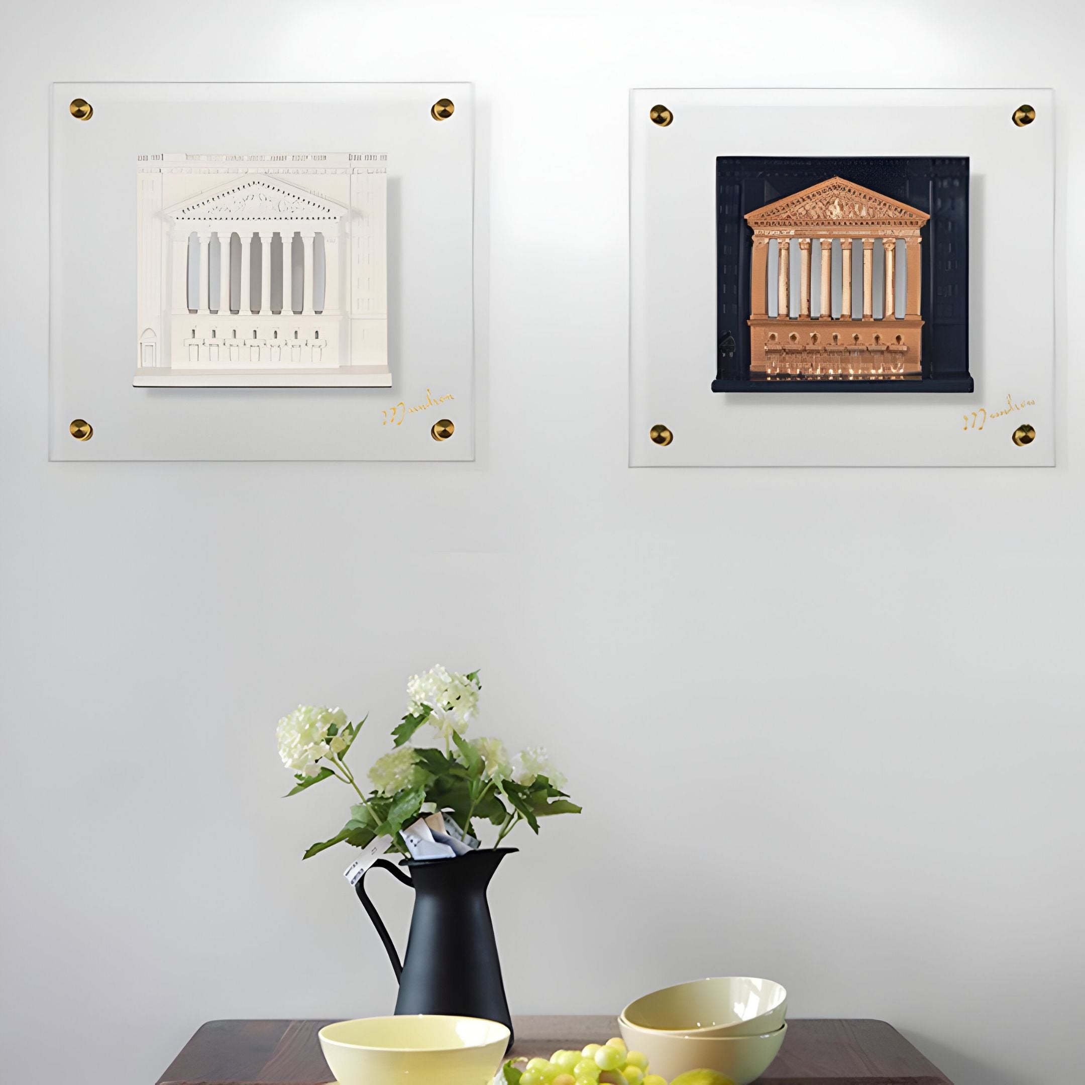 Two framed artworks hang on a white wall above a table set with a black pitcher of white flowers, yellow bowls, and fruit. On the left is the Capitolique White Classical Facade 3D Wall Art by Giant Sculptures; the right piece is brown and black.