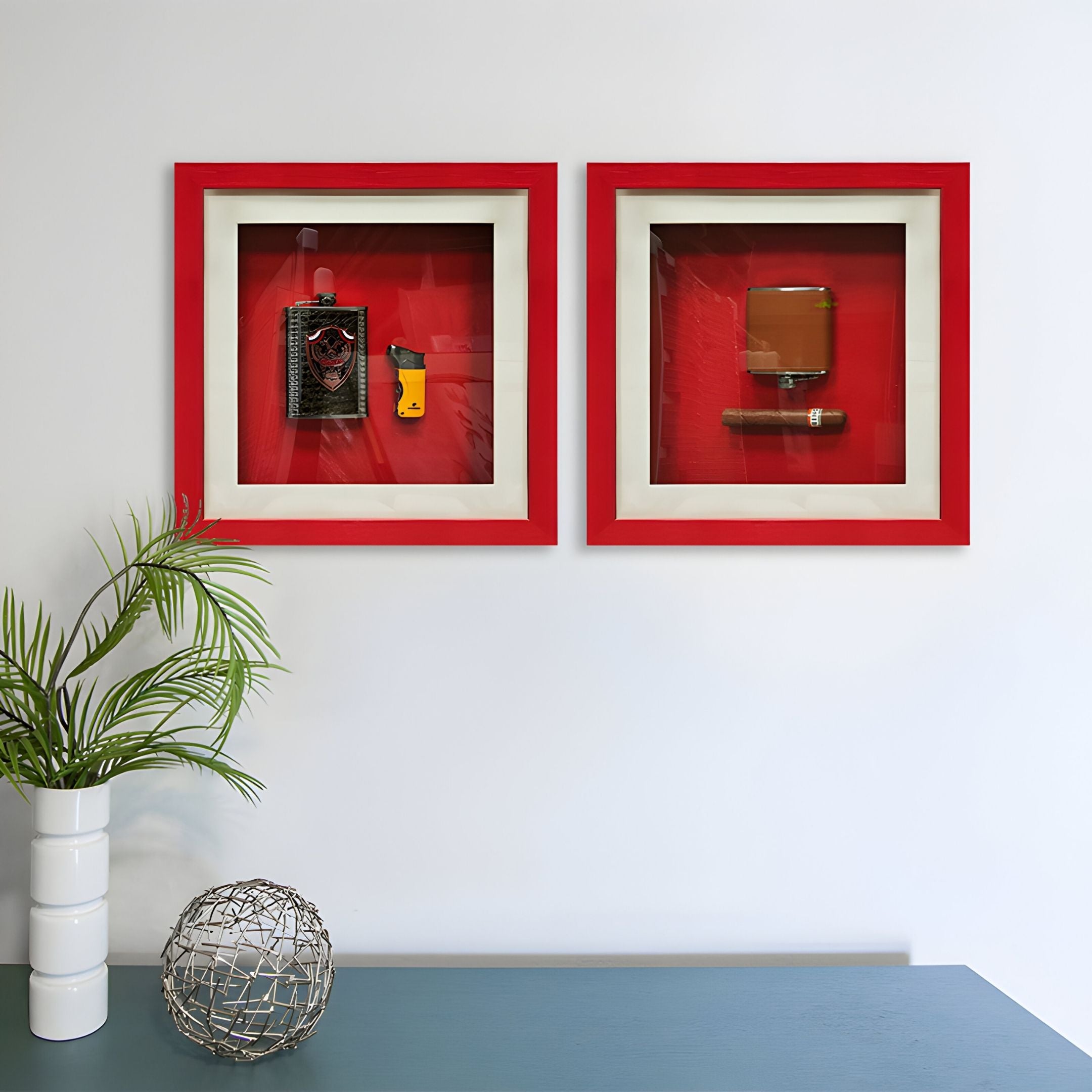 Two Cavendro Flask & Butane Lighter Framed Wall Art pieces by Giant Sculptures hang on a white wall. Below, a green table displays a potted plant and a decorative wire ball.