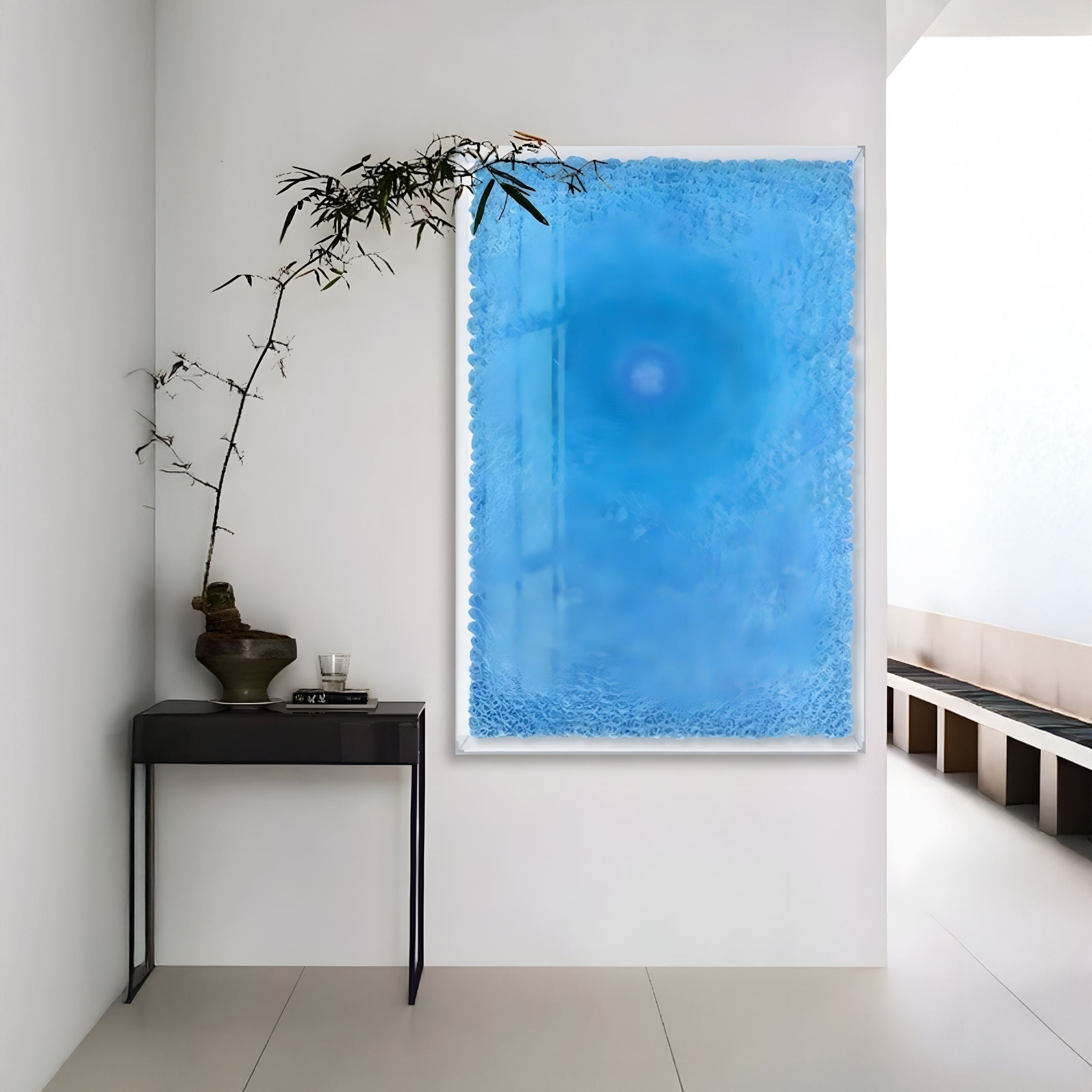 A minimalist room features a small black table with a bonsai and glassware, set against a white wall adorned with Giant Sculptures' Lumora Deep Blue Acrylic Framed 3D Paper Wall Art. Natural light illuminates the neutral decor.