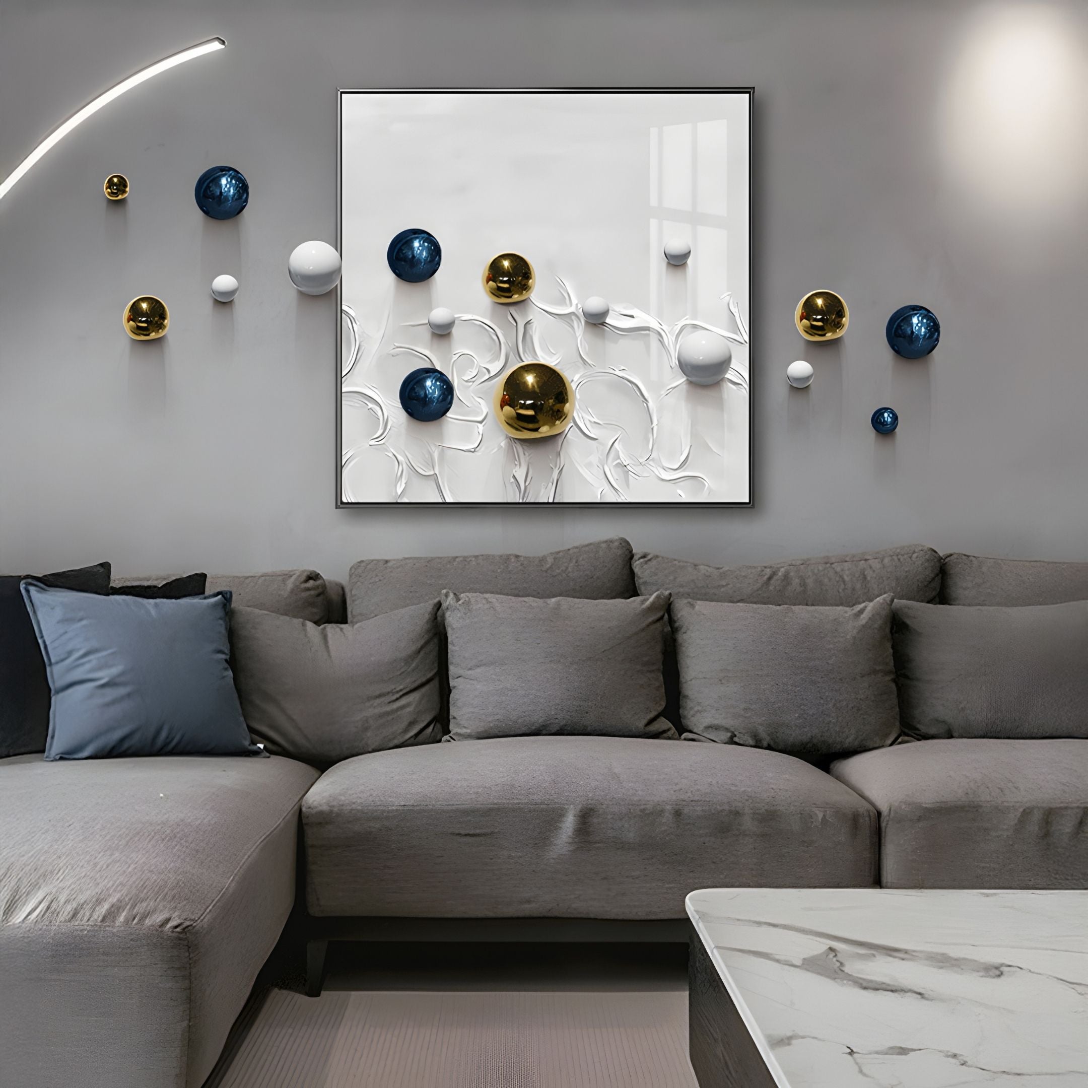 A modern living room features a grey sectional sofa with blue, white, and gold accent pillows. Above it hangs the Aeroluxe White Swirl Abstract 3D Wall Art With Metal Balls by Giant Sculptures, displaying metallic spheres in blue, gold, and white.