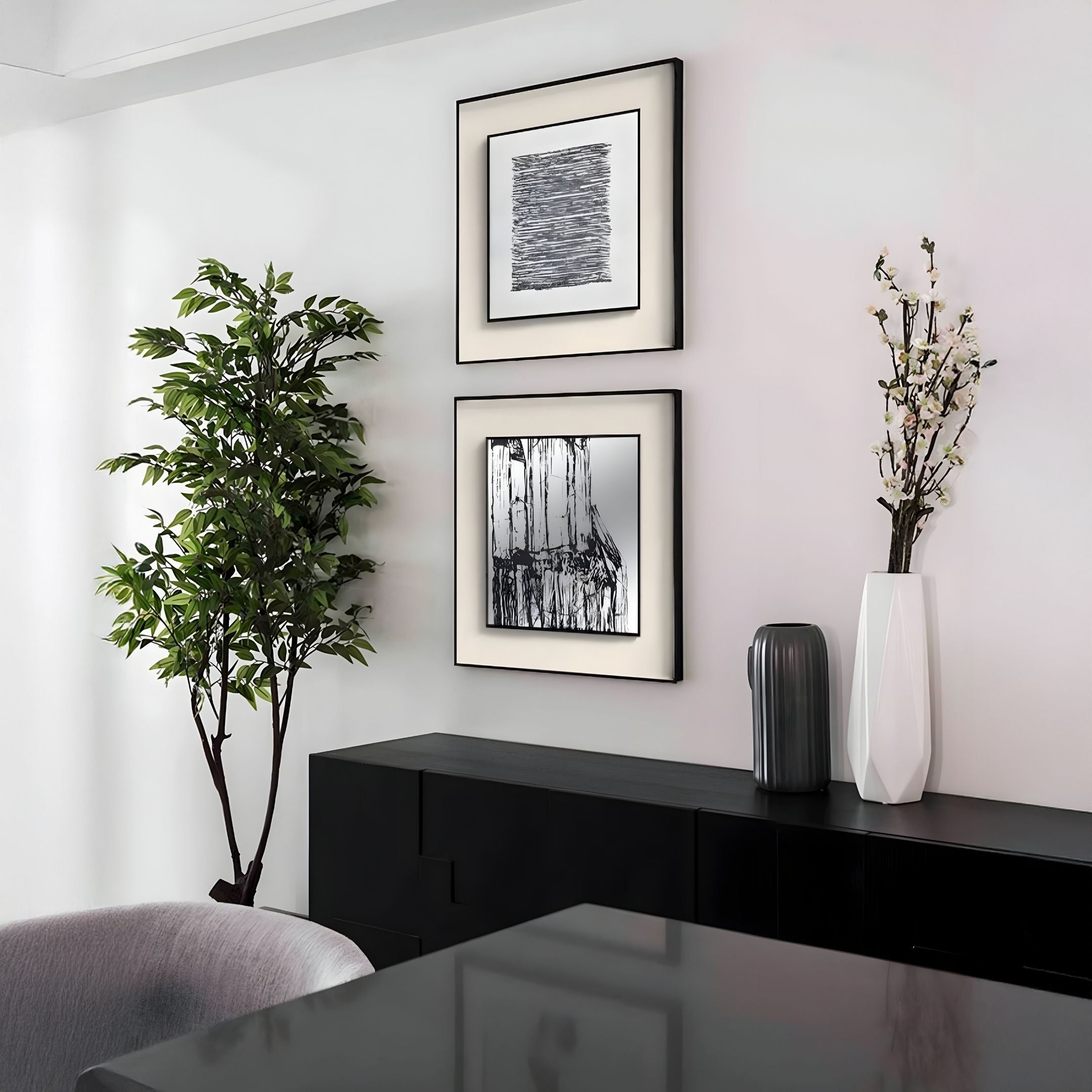 A modern minimalist interior features a black sideboard, decorative vases, a tall leafy plant, and two Stratonis Monochrome Abstract Textured Framed Wall Art pieces by Giant Sculptures on a white wall, with a gray chair and table visible in the foreground.