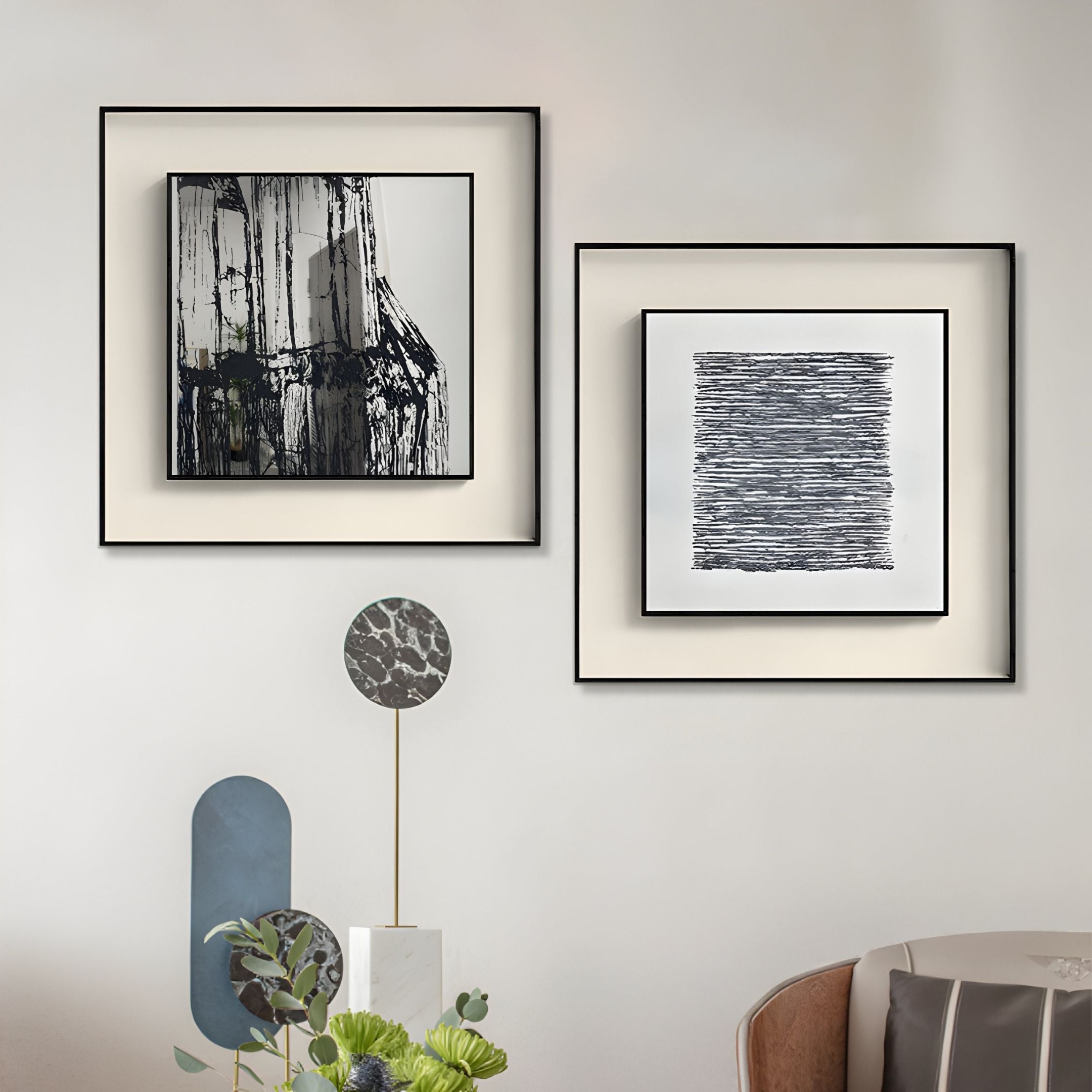 Two Stratonis Grey Linework Textured Framed Wall Art pieces by Giant Sculptures in black frames hang on a light wall above a decorative stand with geometric accents and a green floral arrangement. A beige chair appears in the lower right corner.