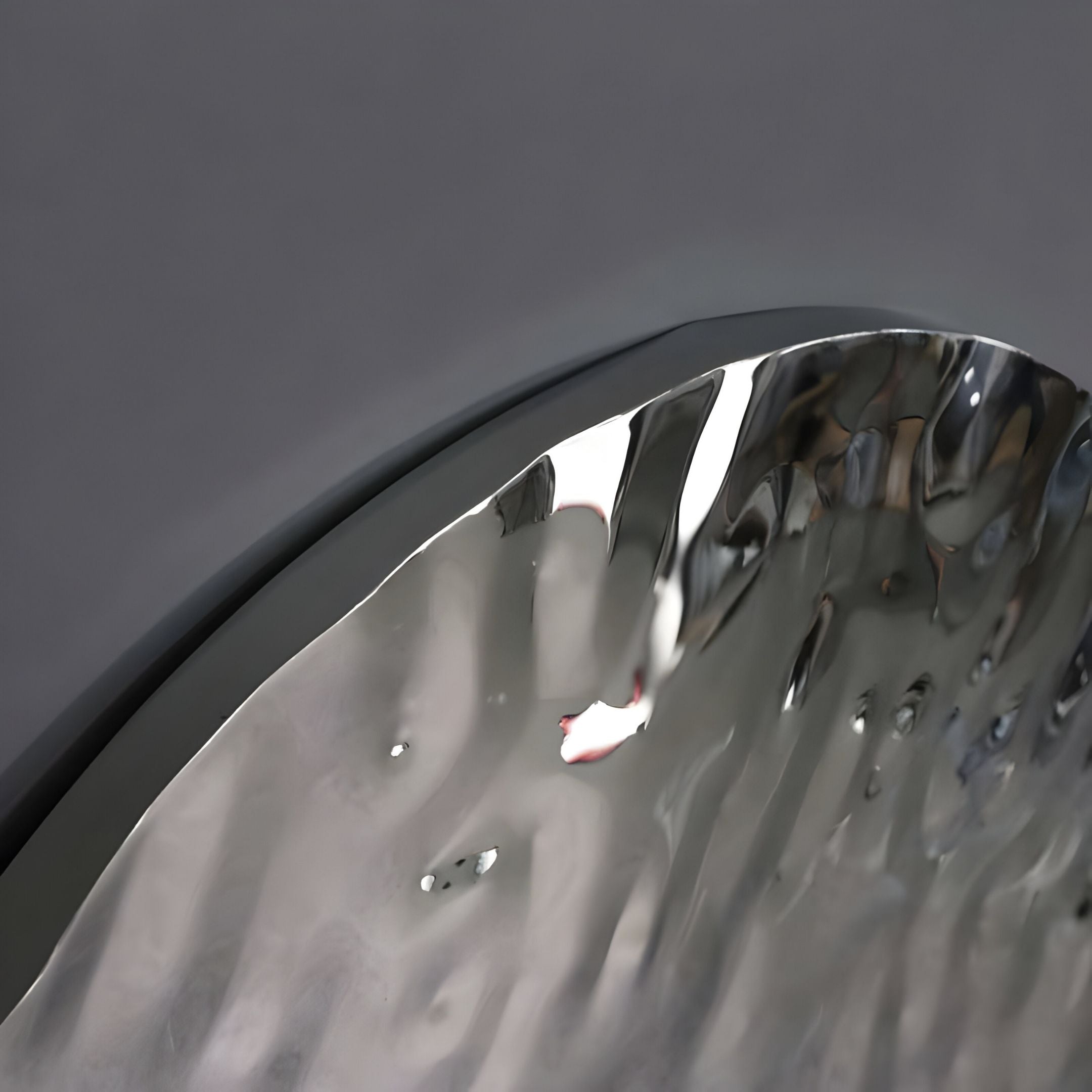 A close-up of the Lumineris Silver Ripple Sphere Metal 3D Wall Art by Giant Sculptures shows its reflective, wavy metallic surface with a curved edge against a smooth gray background, highlighting subtle reflections and artistic distortions.