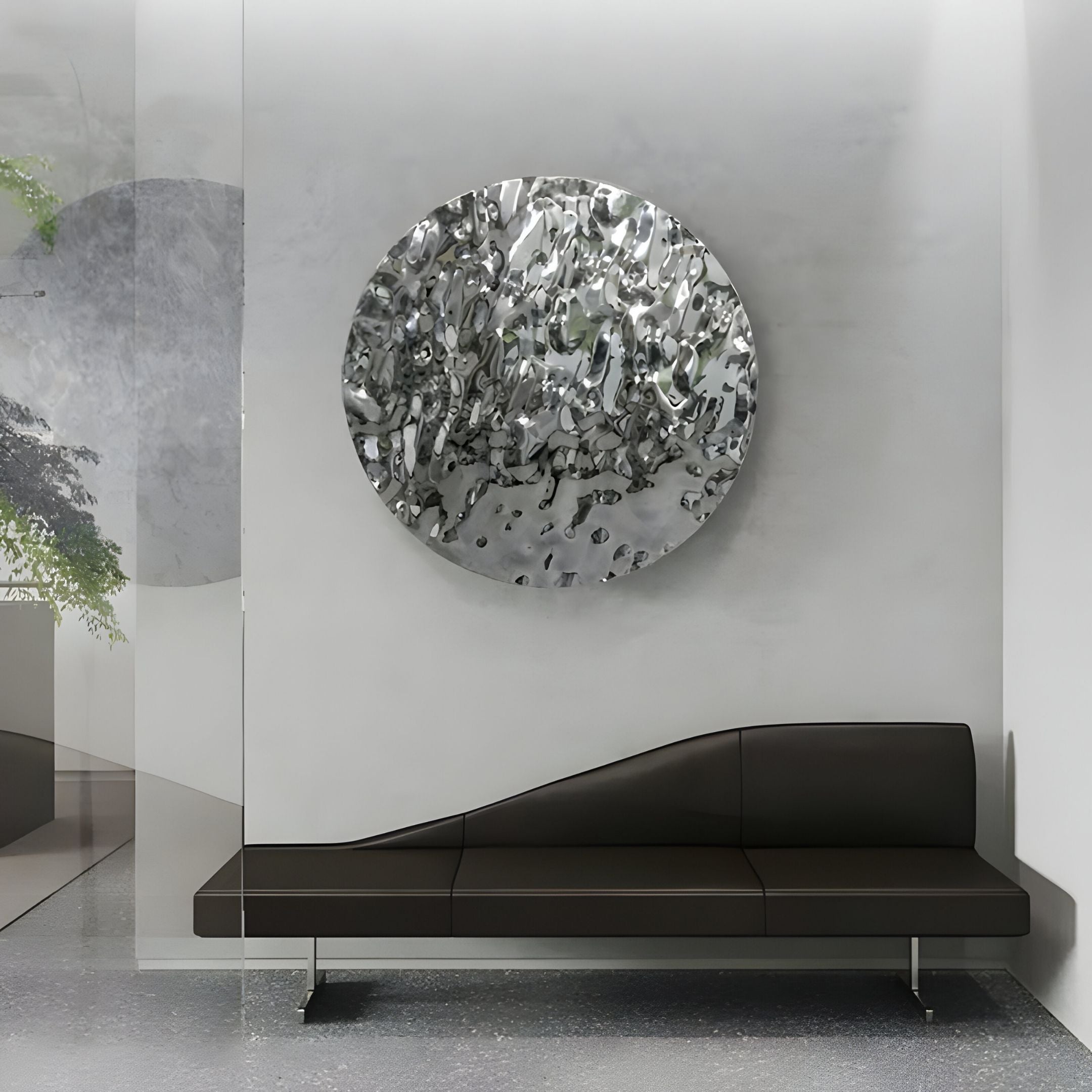 A modern waiting area features a black minimalist bench and the Lumineris Silver Ripple Sphere Metal 3D Wall Art by Giant Sculptures—a striking, round stainless steel piece—mounted on a light gray wall, with a speckled floor and greenery visible through glass.