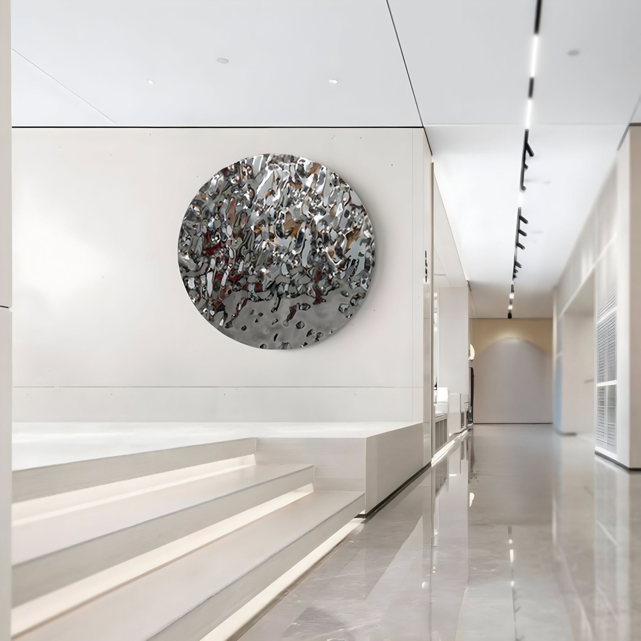 A modern, minimalist interior with glossy floors and white walls showcases the Lumineris Silver Ripple Sphere Metal 3D Wall Art by Giant Sculptures—a captivating piece that reflects its surroundings with a striking silver ripple effect.