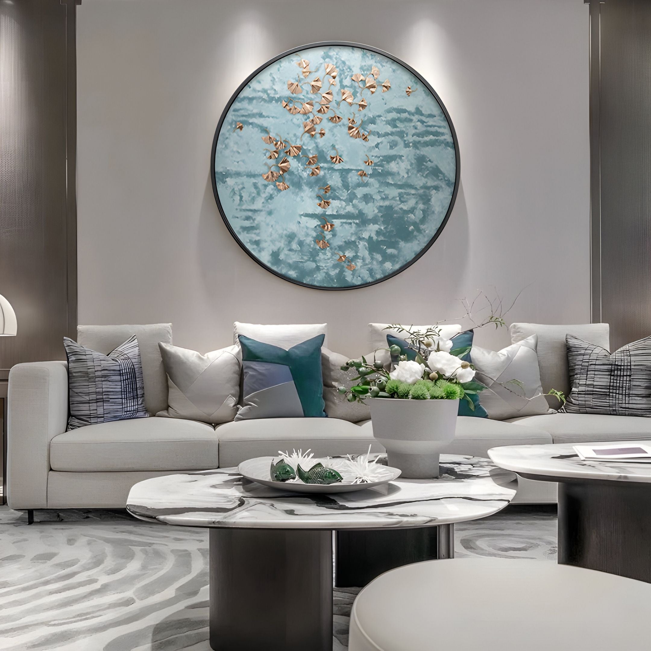 A modern living room with a cream sofa, decorative pillows, marble coffee tables, floral accents, and the Florivonne Gilded Metal Leaf Round 3D Wall Art by Giant Sculptures featuring gold leaves on a textured blue background.