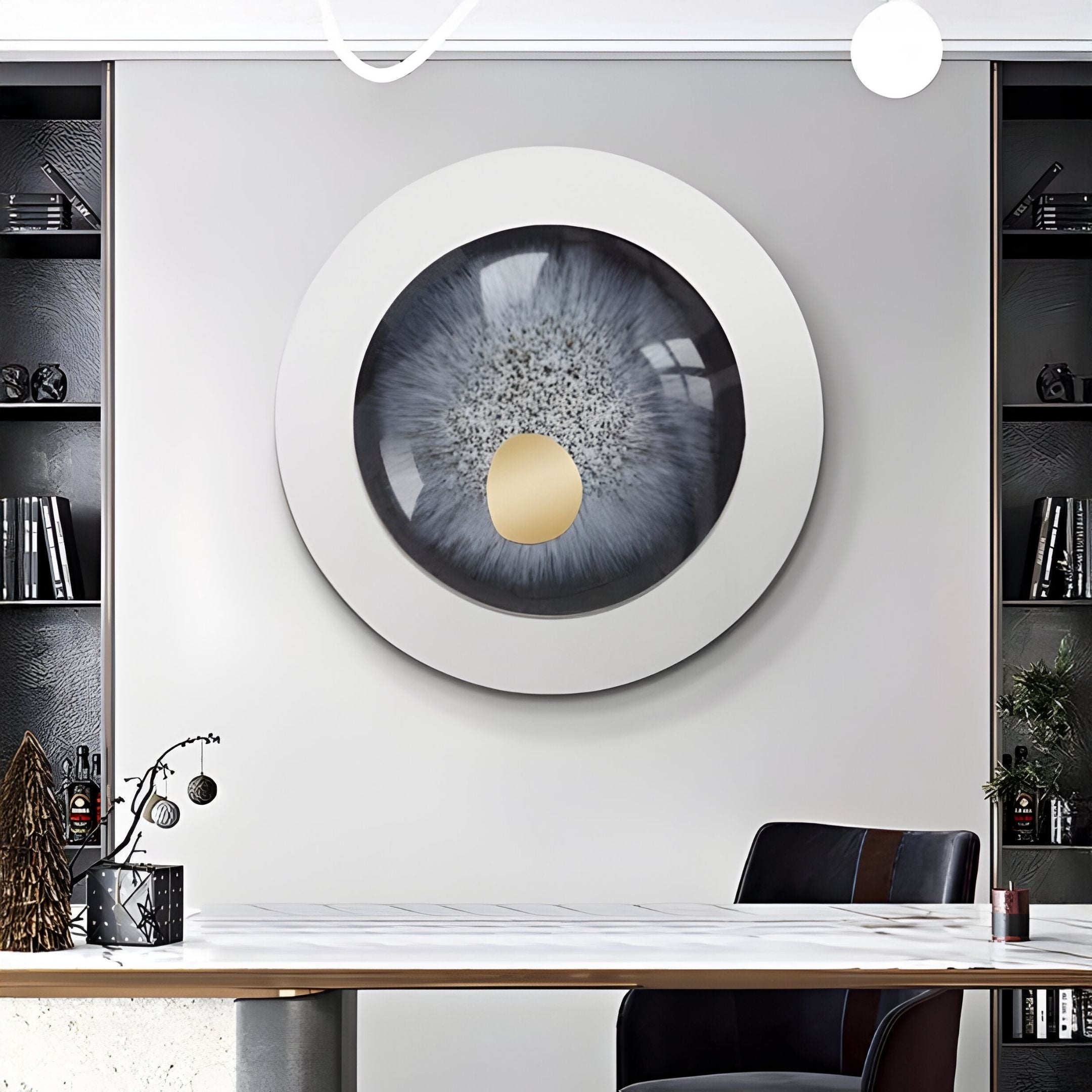 A modern office with shelves and books features the Giant Sculptures Iriselle Frost Grey Crystal Porcelain Round 3D Wall Art—resembling a blue-gray human eye with a gold accent—displayed above a white desk and black chair.