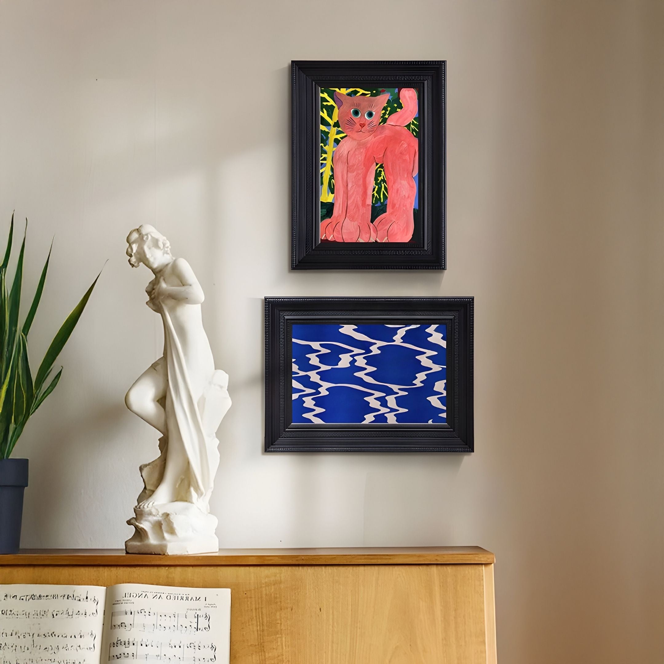 A white woman statue stands on a wooden piano with sheet music. Above, Giant Sculptures’ Ardensir Pink Cat & Blue Ripple 2-Set Framed Wall Art hangs, while a green plant completes the decor.