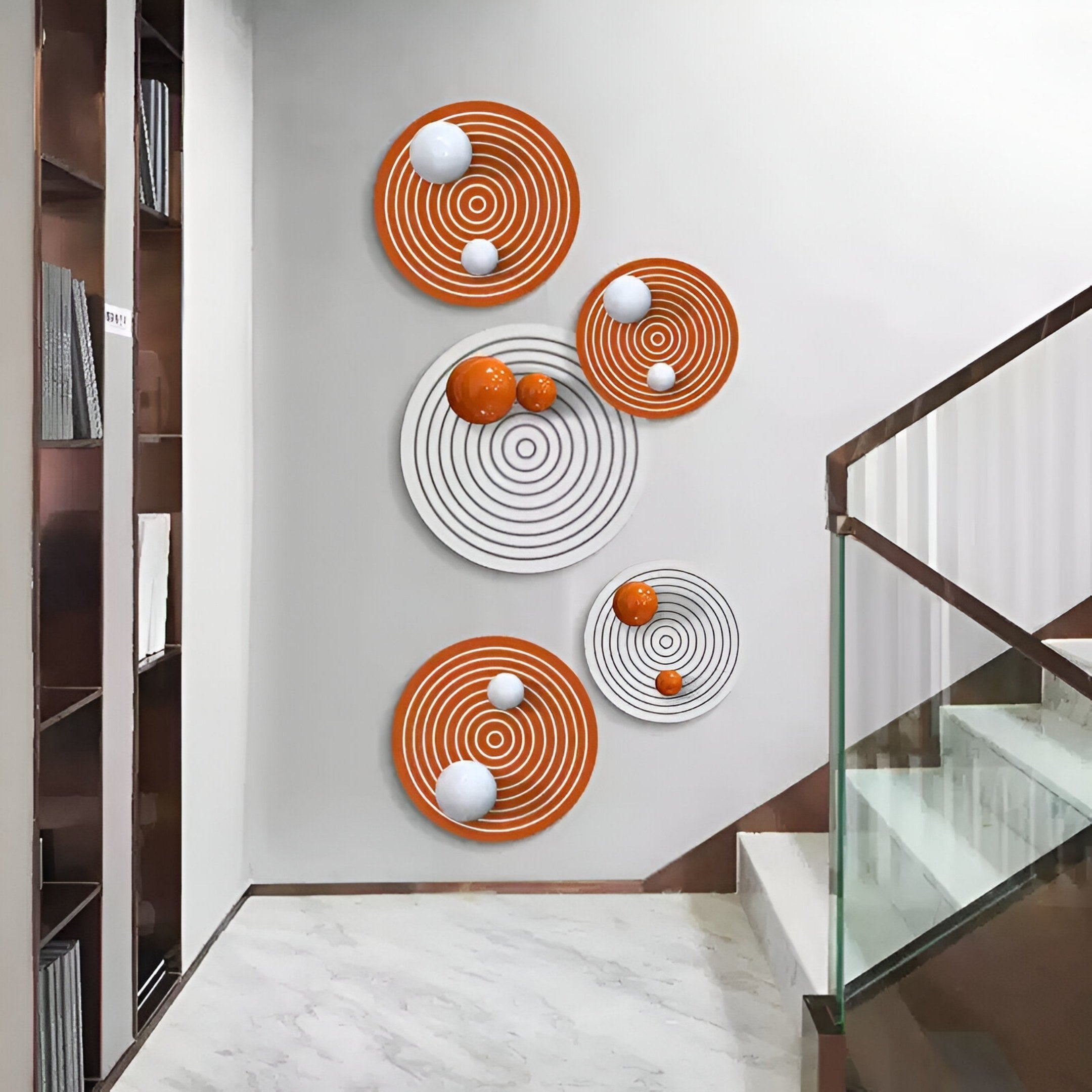 A modern interior wall displays the Orbiora Orange & White Orb 5-Panel Concentric Sphere 3D Wall Art by Giant Sculptures, featuring five circular panels in orange and white beside a staircase with a glass railing and built-in bookshelf.