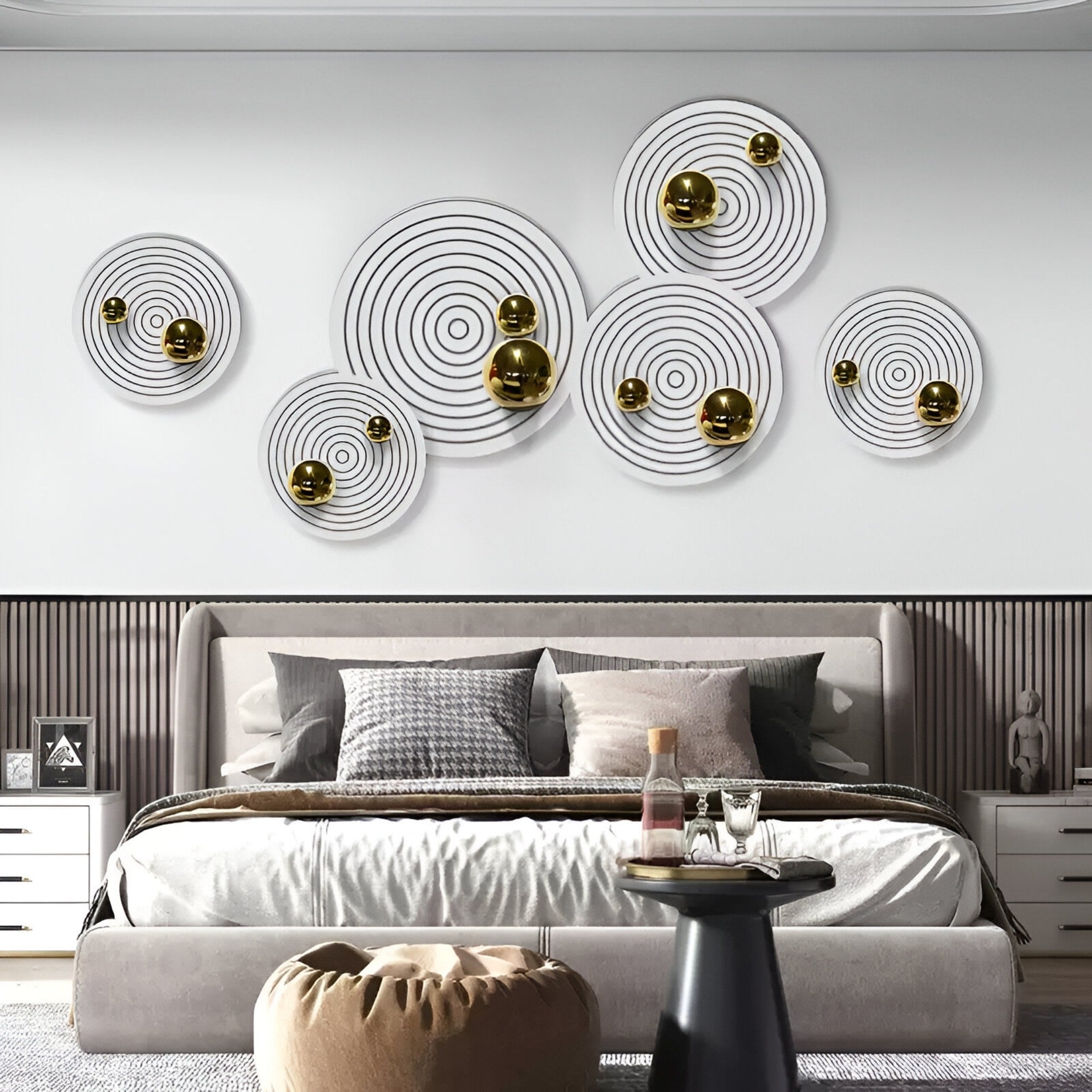A modern bedroom with a bed in neutral bedding, a pouf, and a small round table. Above the bed, Giant Sculptures' Orbiora Gold Orb 6-Panel II Concentric Sphere 3D Wall Art adds sculptural flair with circular forms and gold accents.