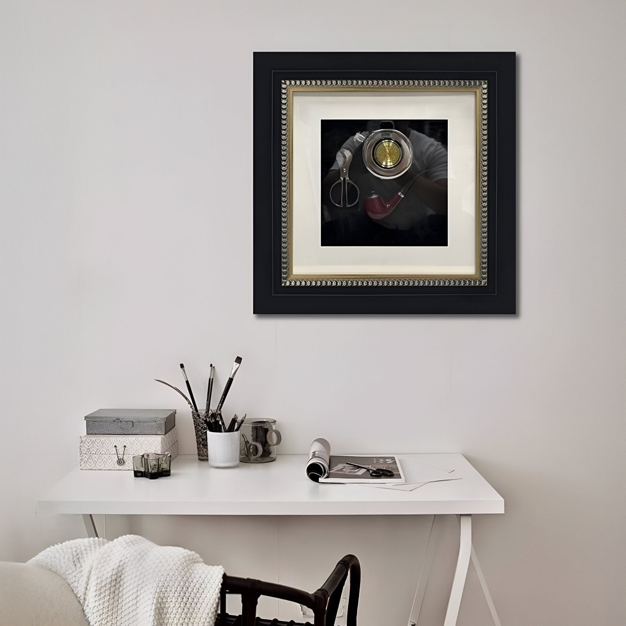 A white desk with art supplies sits against the wall. Above, Giant Sculptures’ Havantra Pipe & Scissors Smokers Set Framed Wall Art brings style, while a chair and decorative boxes create a creative, refined workspace.