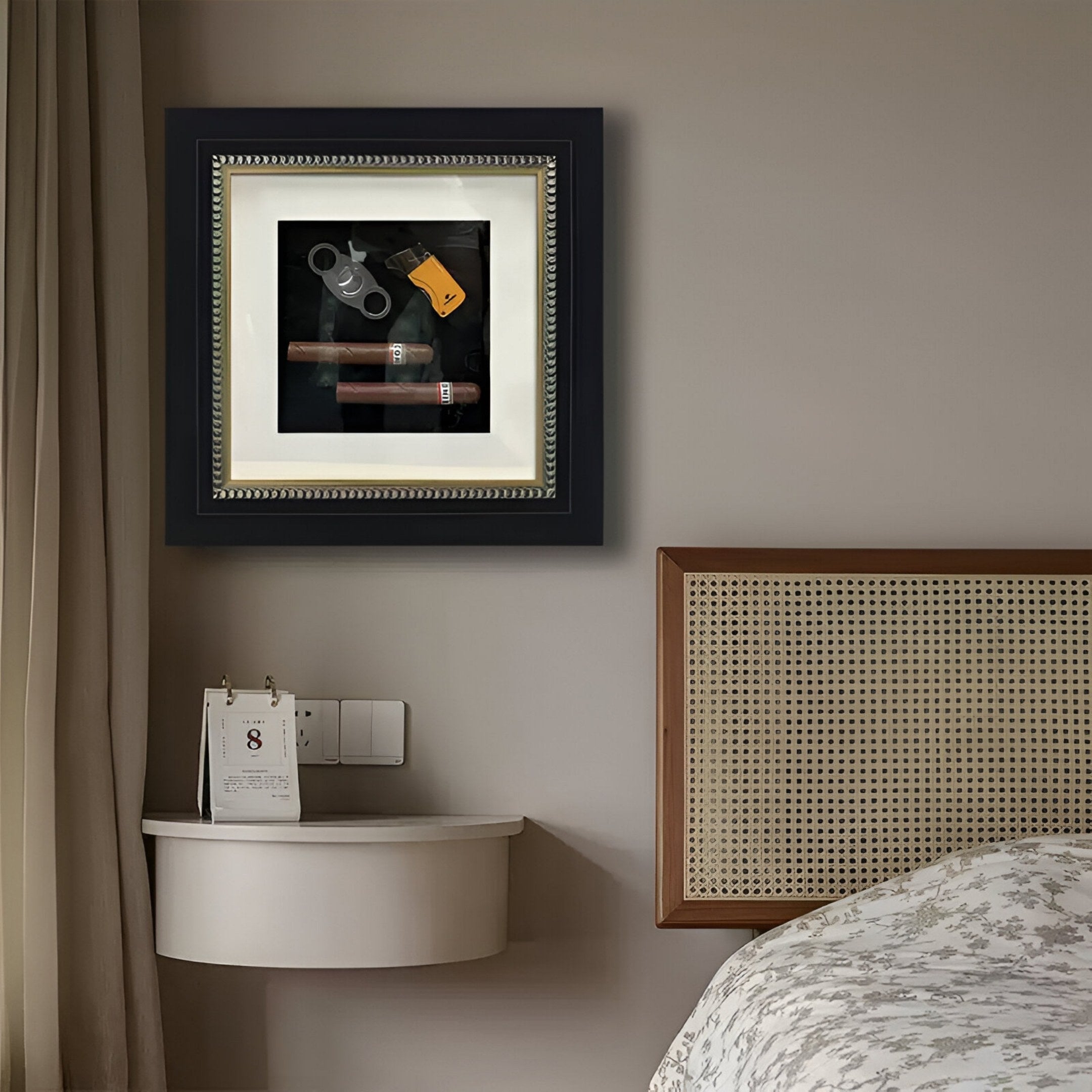 The Havantra Double Cigar & Cutter Framed Wall Art by Giant Sculptures hangs on a beige wall above a floating nightstand with a calendar, beside a bed featuring a rattan headboard.
