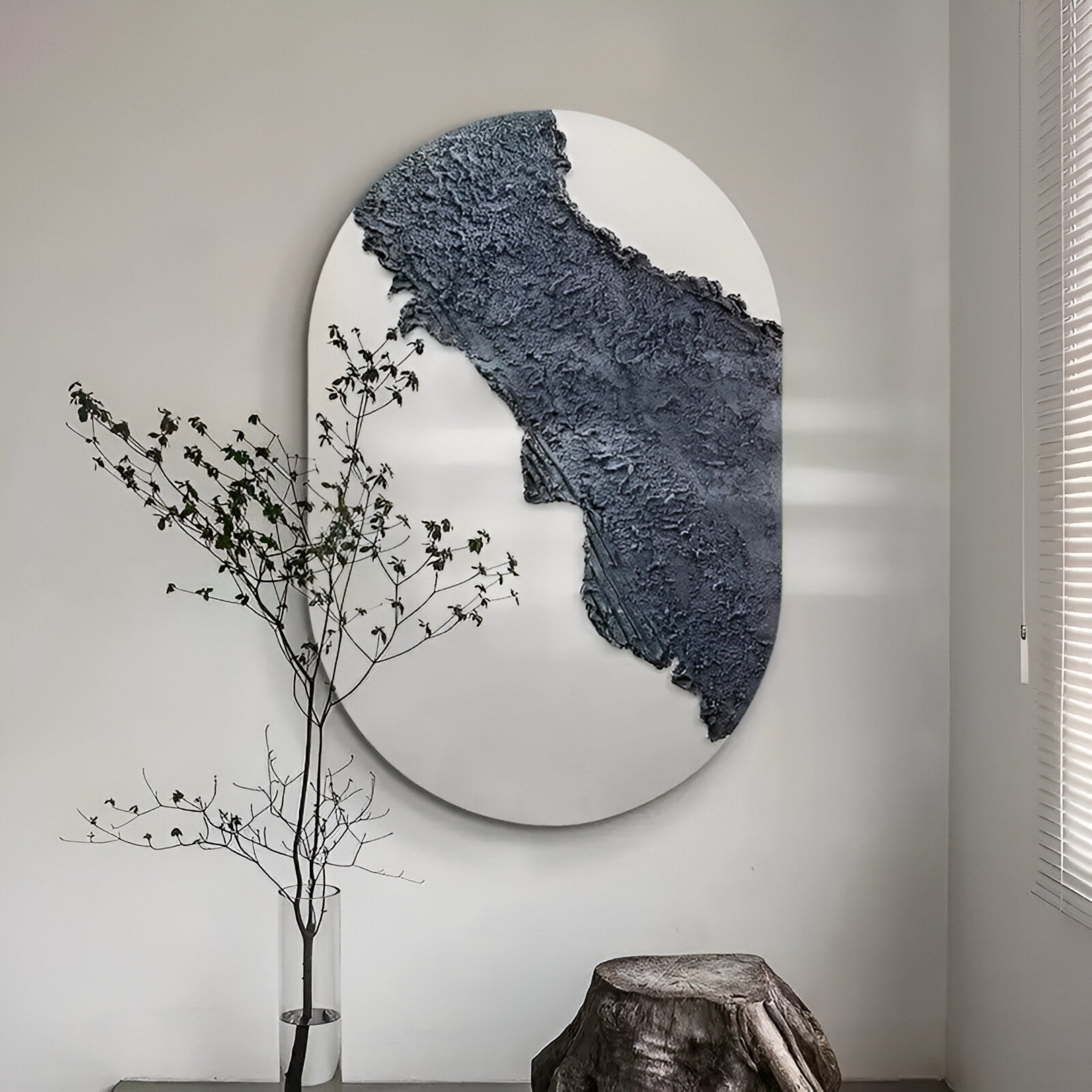 The Orrica Deep Charcoal Textured Relief 3D Wall Art by Giant Sculptures features dark gray texture over a white oval. A minimalist branch in a glass vase and a rustic tree stump table sit nearby, illuminated by sunlight through blinds.