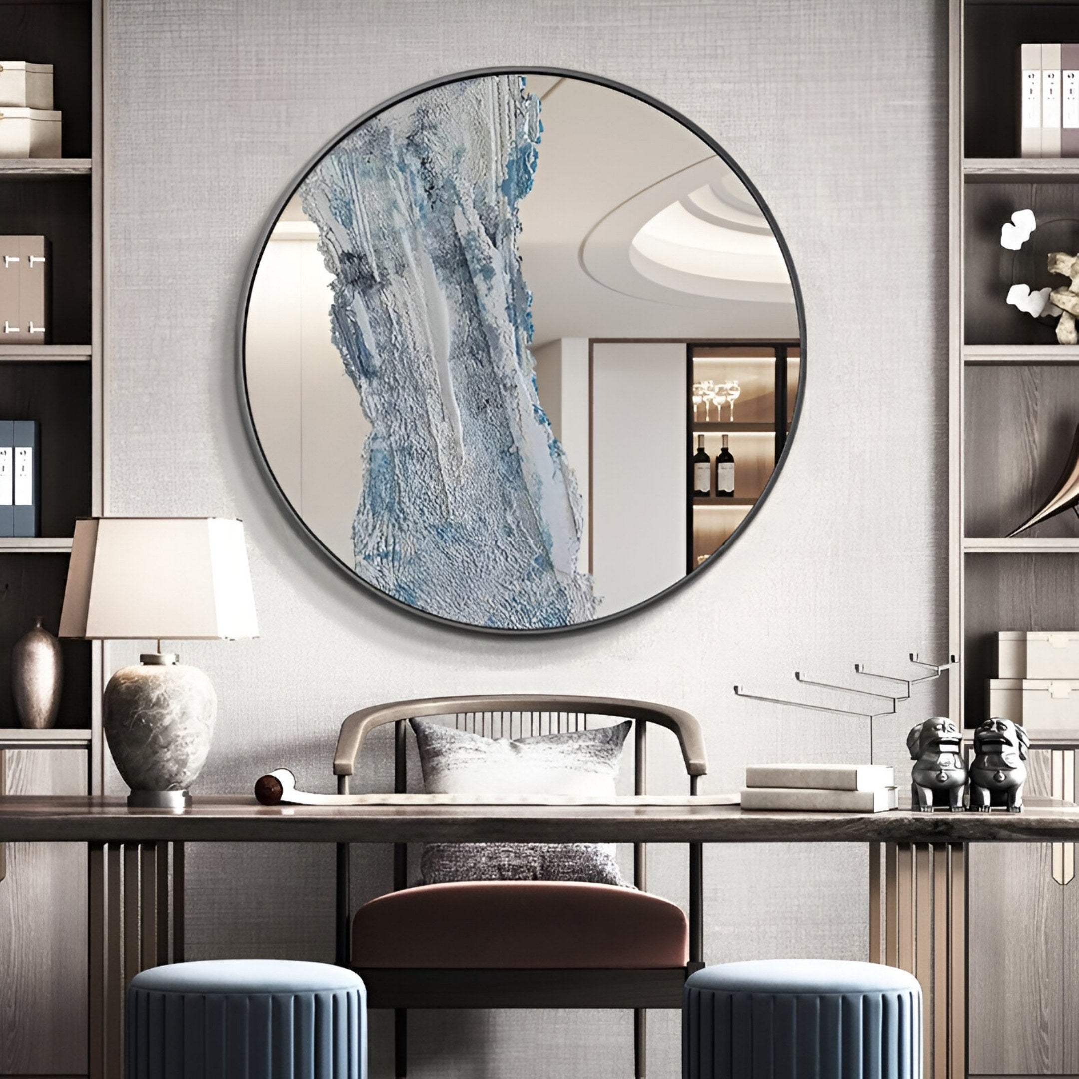A modern study features shelves, a desk with the Erosian Silver Cascade Textured Round Mirror 3D Wall Art by Giant Sculptures above it, a table lamp, decorative objects, a velvet chair, and two blue stools for an elegant workspace.