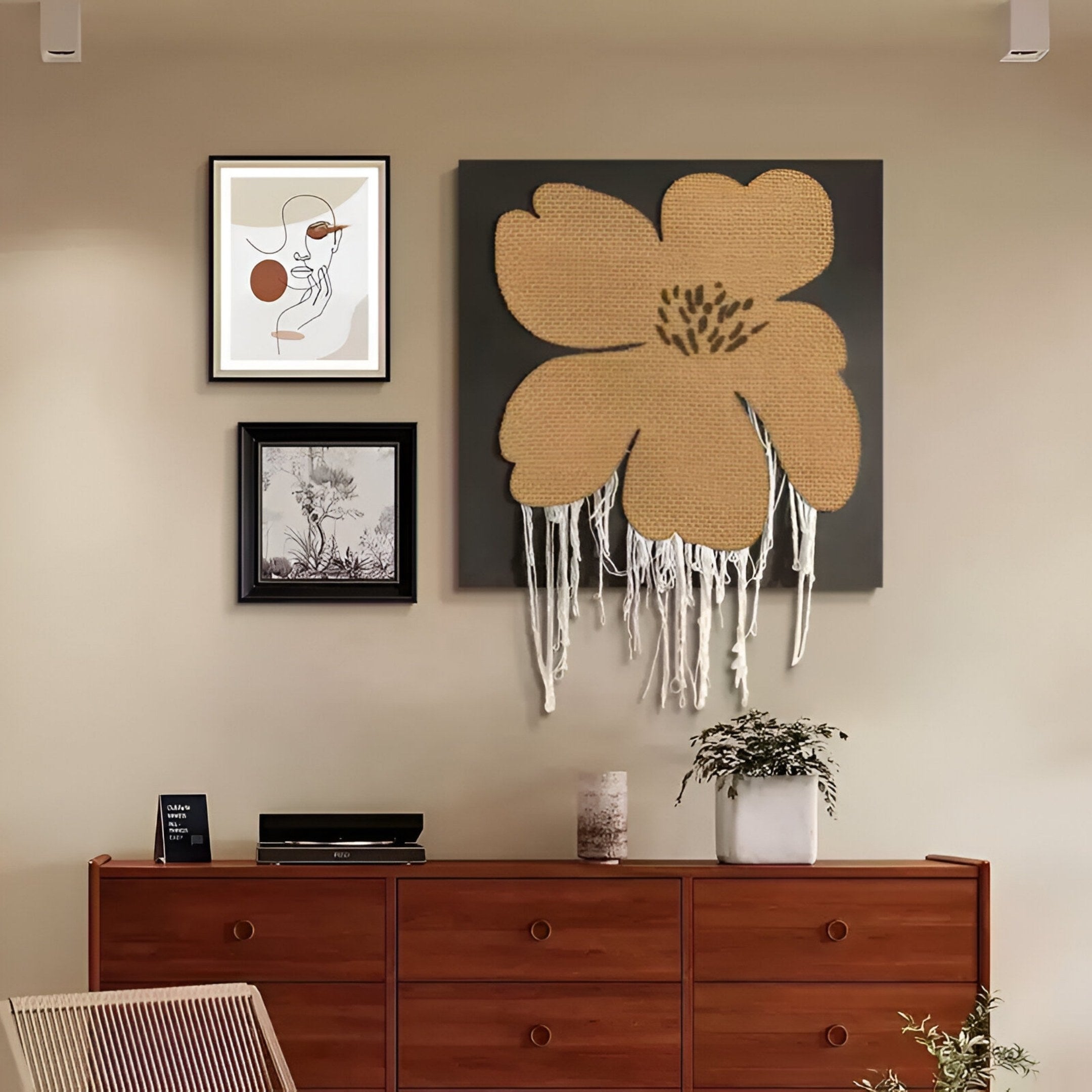 A wooden dresser topped with a plant sits against a beige wall. Above, the Florayne 3-Set Ochre Woven Textured 3D Wall Art by Giant Sculptures features an abstract face, black and white landscape, and yellow flower with white threads.