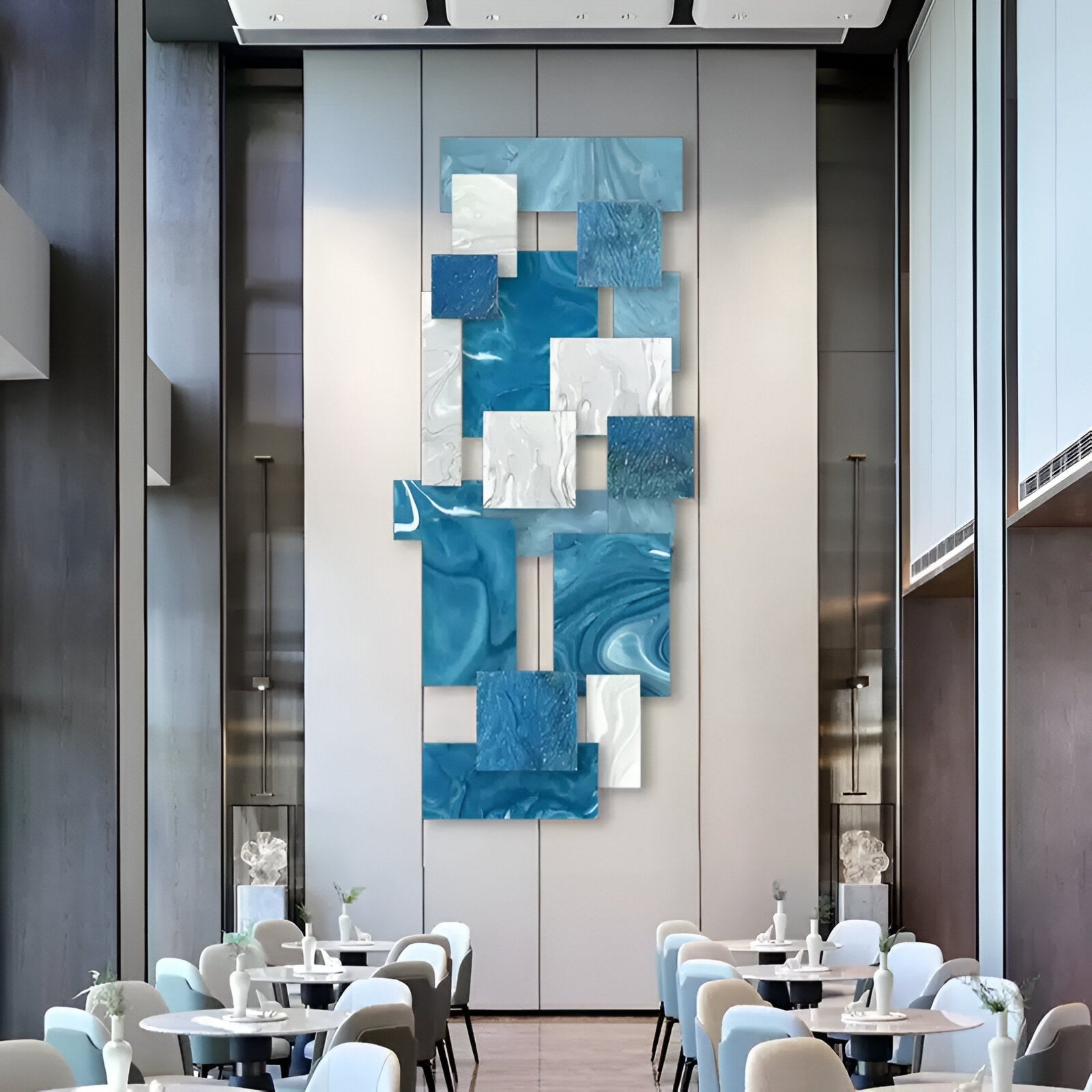 A modern dining area with light chairs and tables showcases the Stravane Blue & White Mosaic Oil Canvas 3D Wall Art by Giant Sculptures—an abstract installation of vertical rectangular panels on a tall, light gray wall.