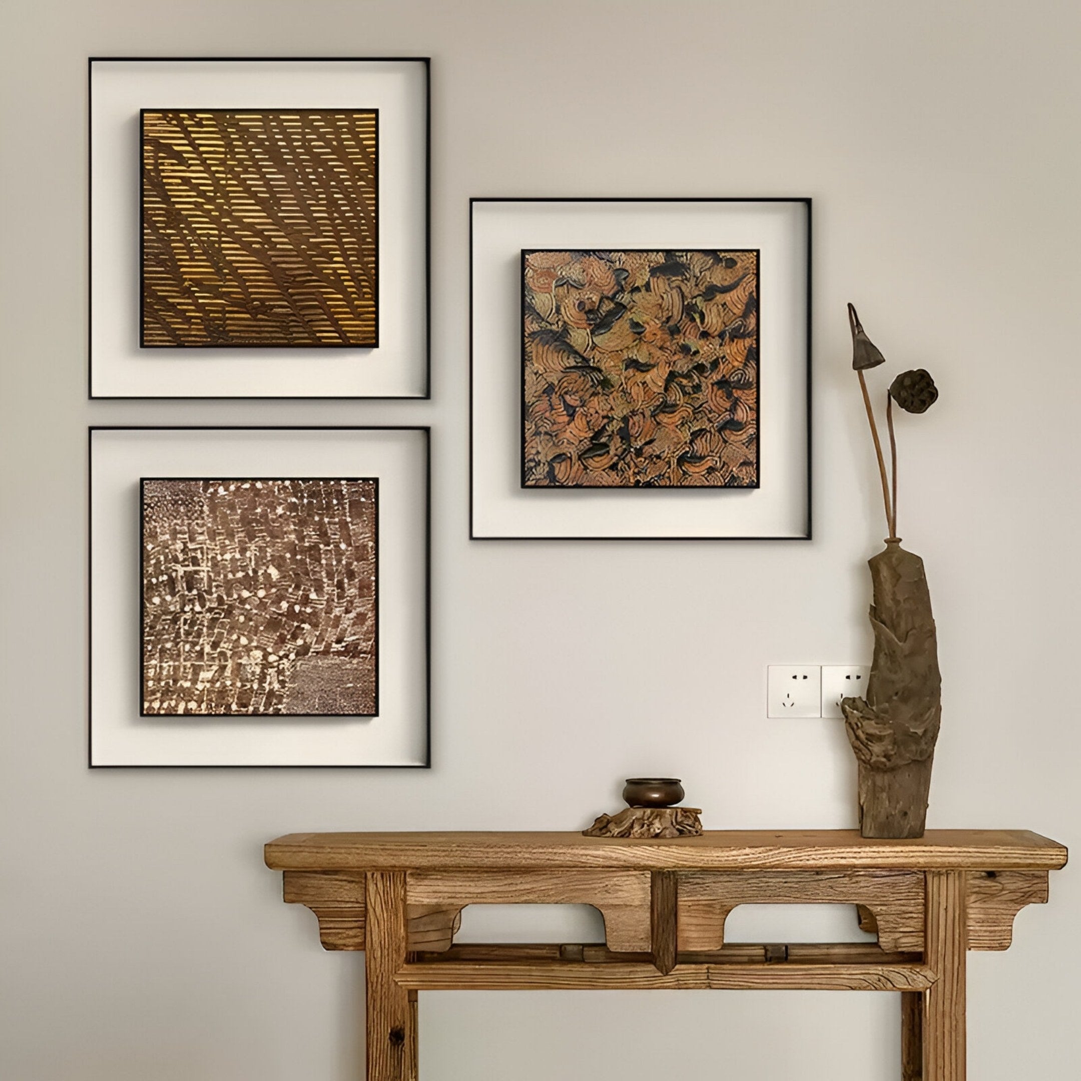 A wooden console table with a small bowl and tall sculpture stands against a beige wall, above which three Fiorenze Bronze Textured Square 3D Wall Art pieces by Giant Sculptures are neatly arranged.