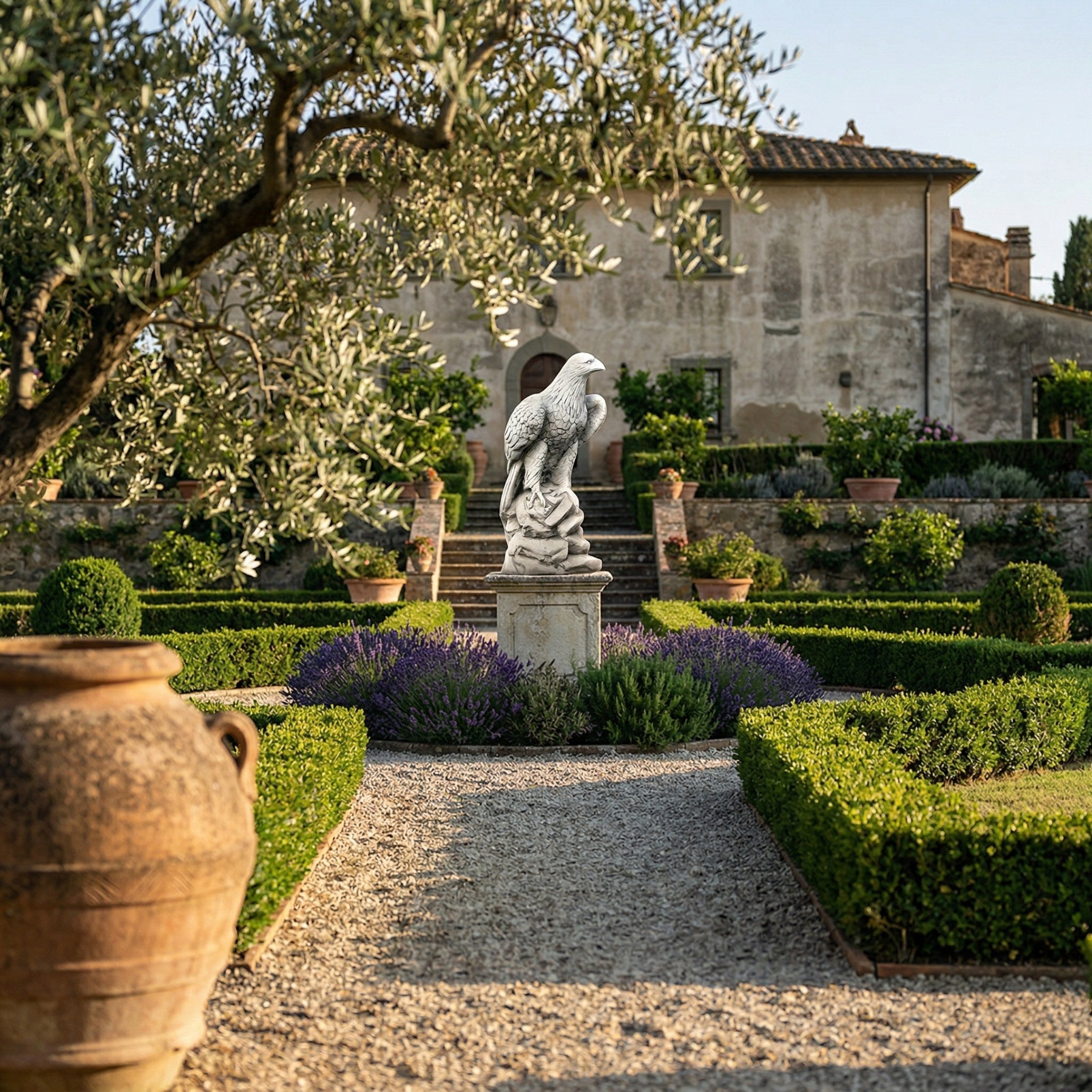A gravel path leads to the Giant Sculptures Regal Eagle White Marble Outdoor Sculpture (120cm), set among trimmed hedges and purple flowers in front of a stone villa with potted plants and olive trees under a clear sky.