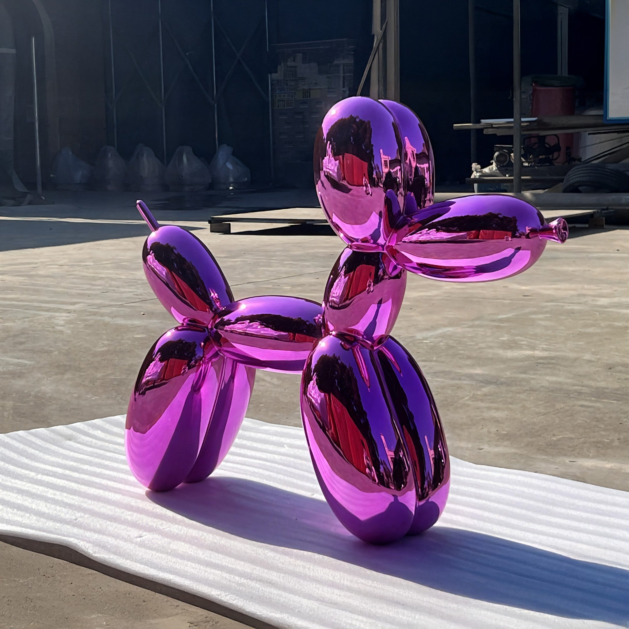 The Flamingo Pink Balloon Dog Sculpture - 80cm by Giant Sculptures shines on a white mat outdoors, reflecting light and its setting—an eye-catching modern centerpiece ideal for stylish decor.
