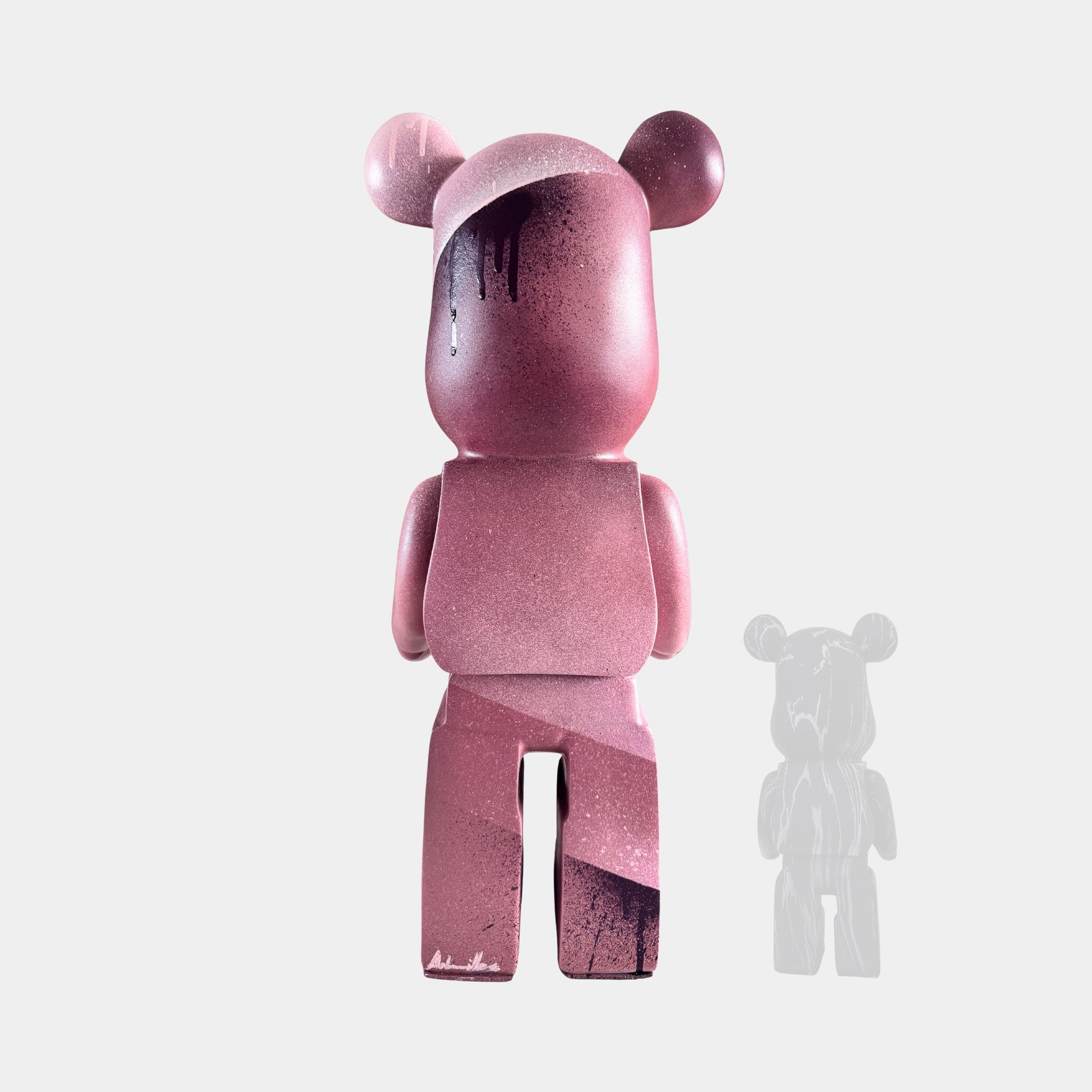 The Giant Sculptures Pinque Bear Sculpture - 50cm stands upright in pink with dark and light paint splatters and a black drip on its head. A smaller, faded grayscale version of this limited edition art toy appears in the background.