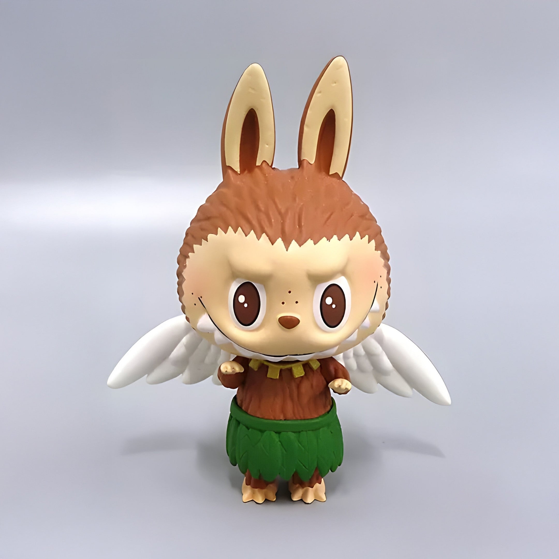 The Space Adventures Zimomo Beast Labubu Sculpture - 100cm by Giant Sculptures features a round, smiling face, rabbit-like ears, brown fur, white wings, and a green grass skirt against a light gray background.