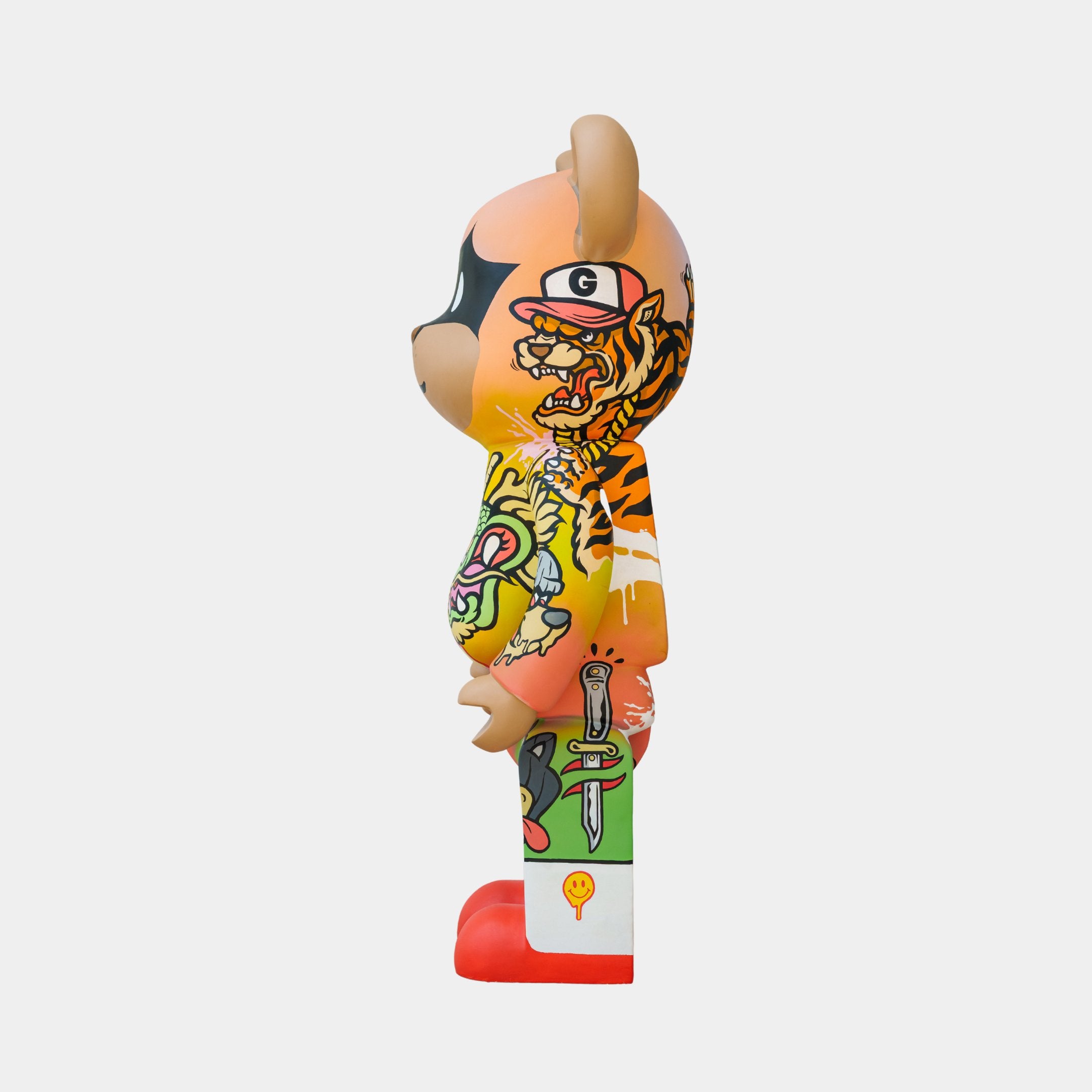 The limited edition Lucky Paws Bear Sculpture by Giant Sculptures is a 50cm artwork featuring a vibrant, graffiti-style bear with tiger, dagger, and heart motifs. It has a brown head and an orange and green body with red feet on a white background.