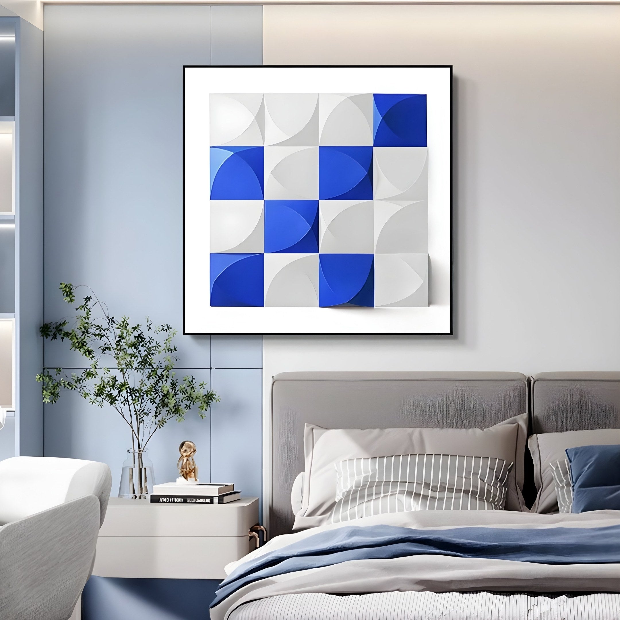 A modern bedroom displays a bed, pillows, and a nightstand with books and a plant. The Geometrica Bold Blue & White Grid 3D Wood Relief Wall Art from Giant Sculptures decorates the light blue wall, adding depth and elegance to the serene space.