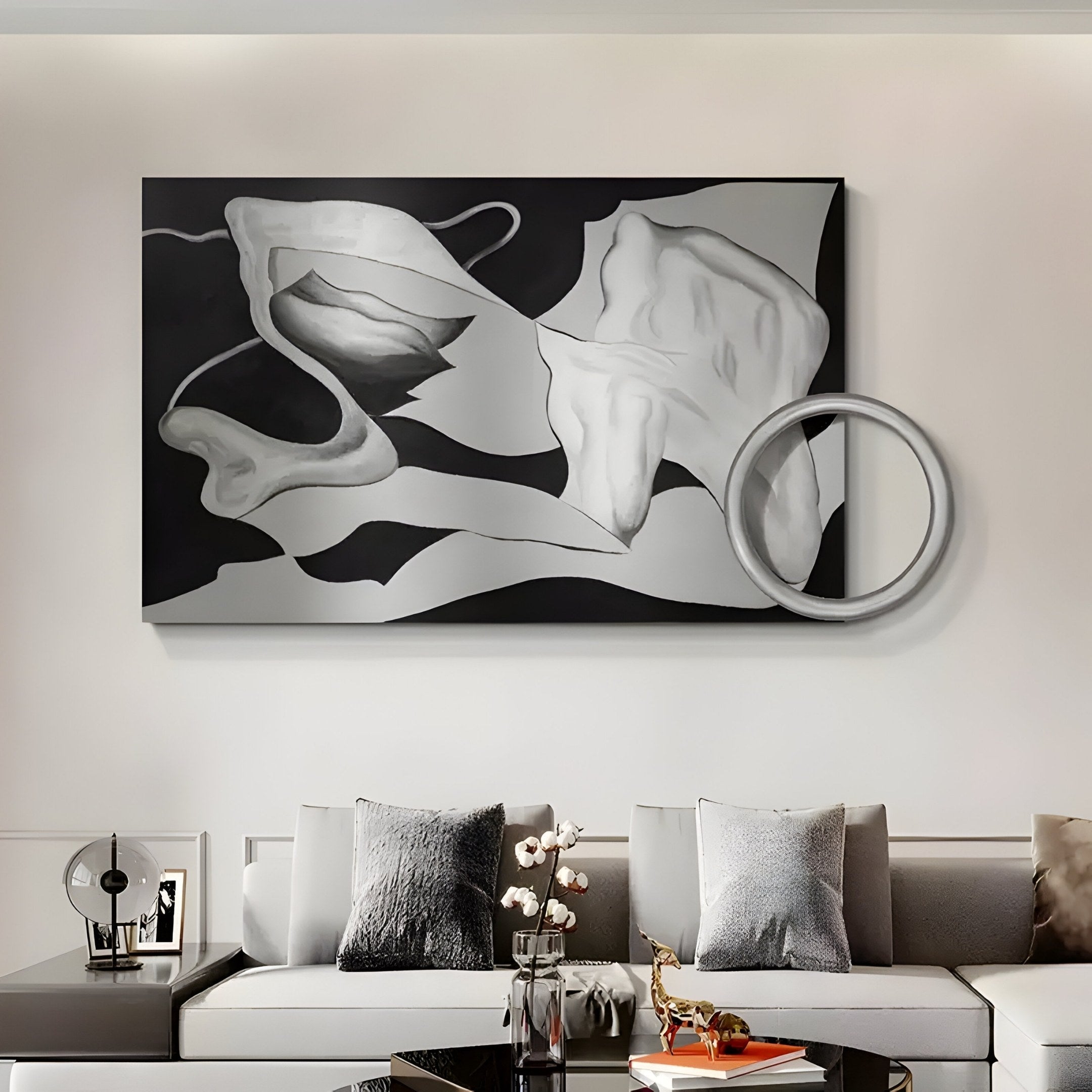 The Calmant Black & Silver Abstract Hand-Painted Wall Art by Giant Sculptures hangs above a modern gray sofa with assorted cushions. A small table in front displays flowers and a gold giraffe statue, adding charm with a touch of wabi-sabi aesthetic.