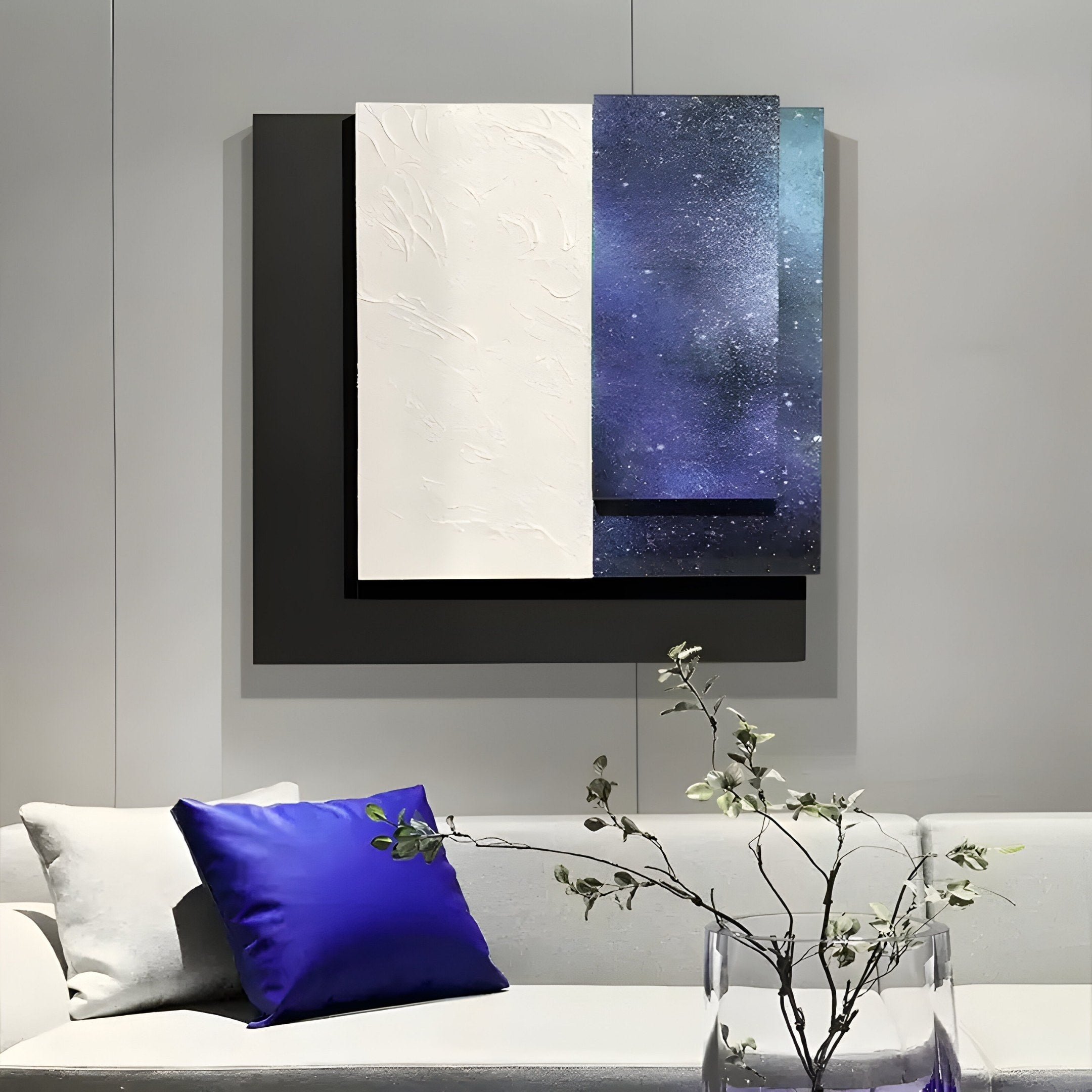 The Astra Galaxy Blue Textured Wood Carving 3D Wall Art from Giant Sculptures graces the wall with its celestial and geometric design of layered black, white, and starry purple-blue rectangles. Below it, a white sofa with a vibrant blue pillow sits beside a glass vase containing delicate flowering branches.