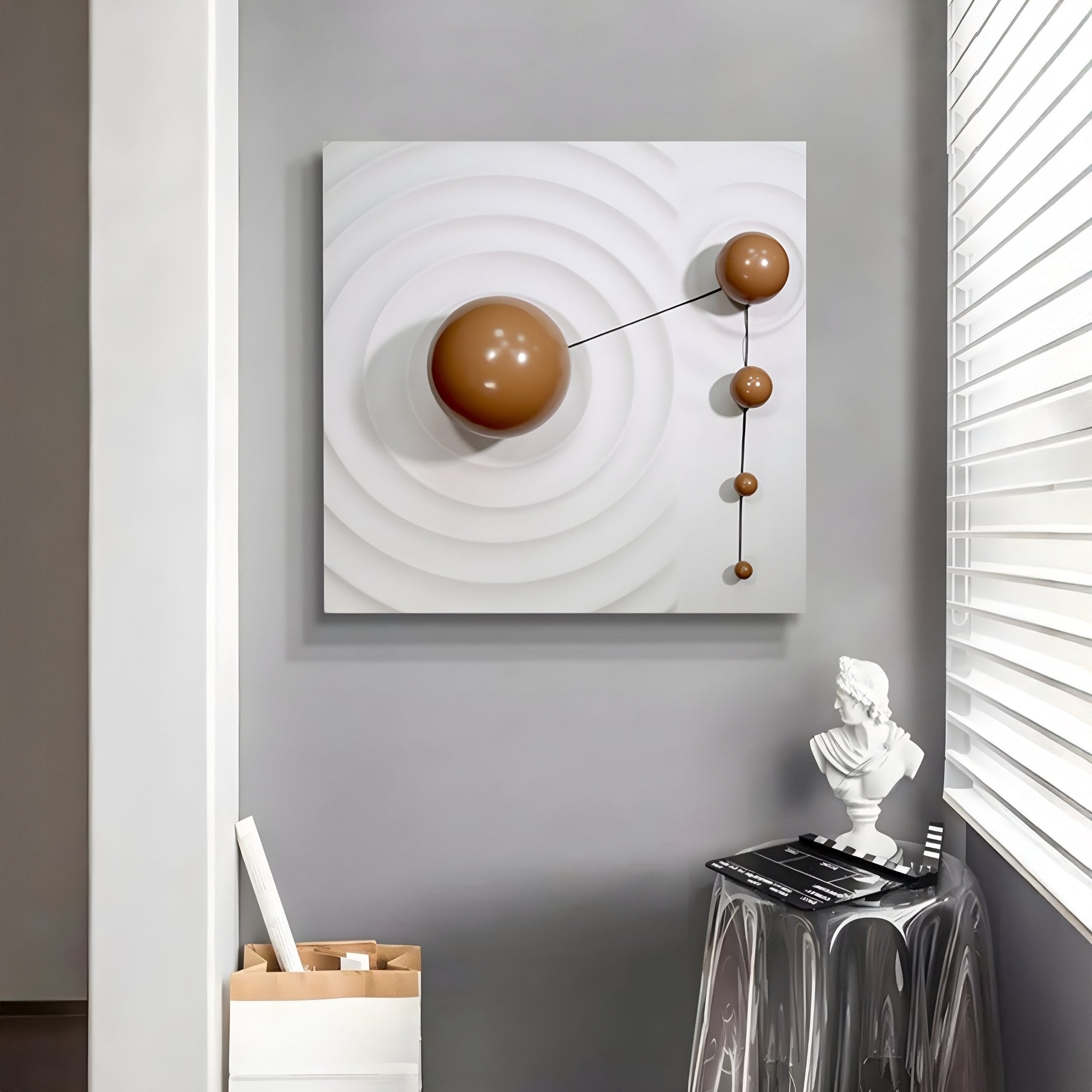 The Galactica White Wood Relief 3D Wall Art by Giant Sculptures features three large brown metal spheres connected by thin rods against concentric circles on a white background. A small white bust statue and a transparent chair are placed near a window with closed blinds.