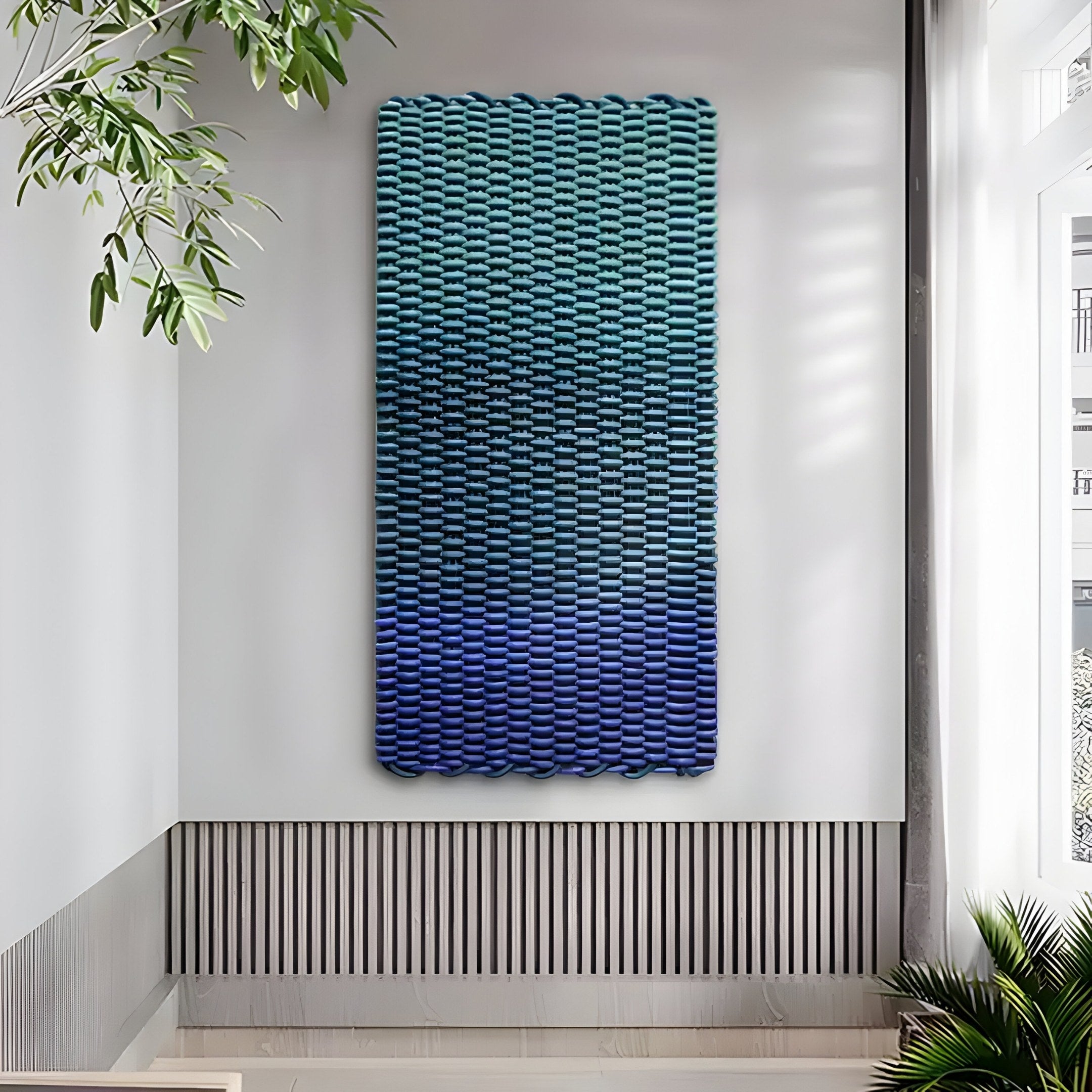 The Spectrum Weave Vertical Midnight Blue Wood Relief 3D Wall Art by Giant Sculptures, featuring a textured, wavy surface in gradient blue and teal shades, hangs on a light gray wall. The minimalist room is enhanced by large windows and potted plants adding greenery.