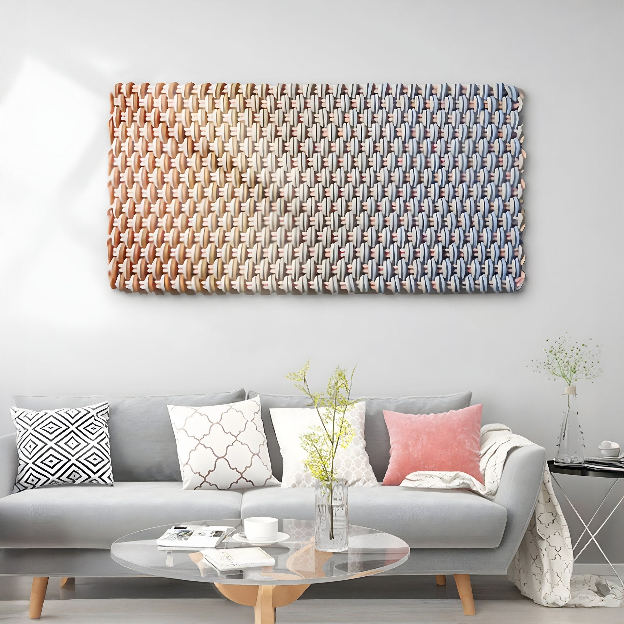 A modern living room includes a gray sofa with black, white, and pink pillows. In front sits a circular glass coffee table topped with flowers in a vase. The wall features Giant Sculptures Spectrum Weave Horizontal Earthy Ombre Wood Relief 3D Wall Art, adding natural warmth to the space.