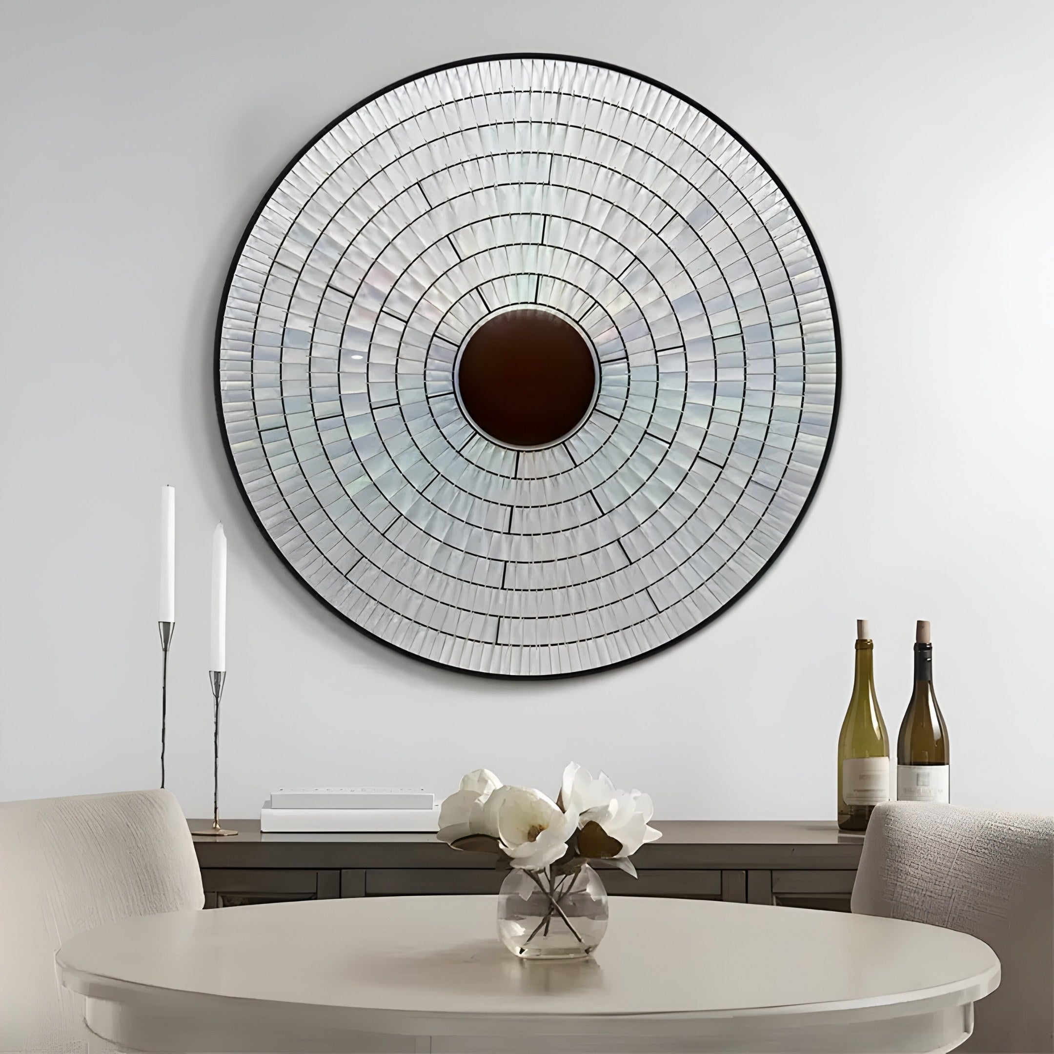 The Solis Radiant Deep Red Core Wood Relief 3D Wall Art by Giant Sculptures is displayed above a console table holding two unlit white candles, wine bottles, and a white vase with flowers. In the foreground, theres a round table and a light-colored chair.