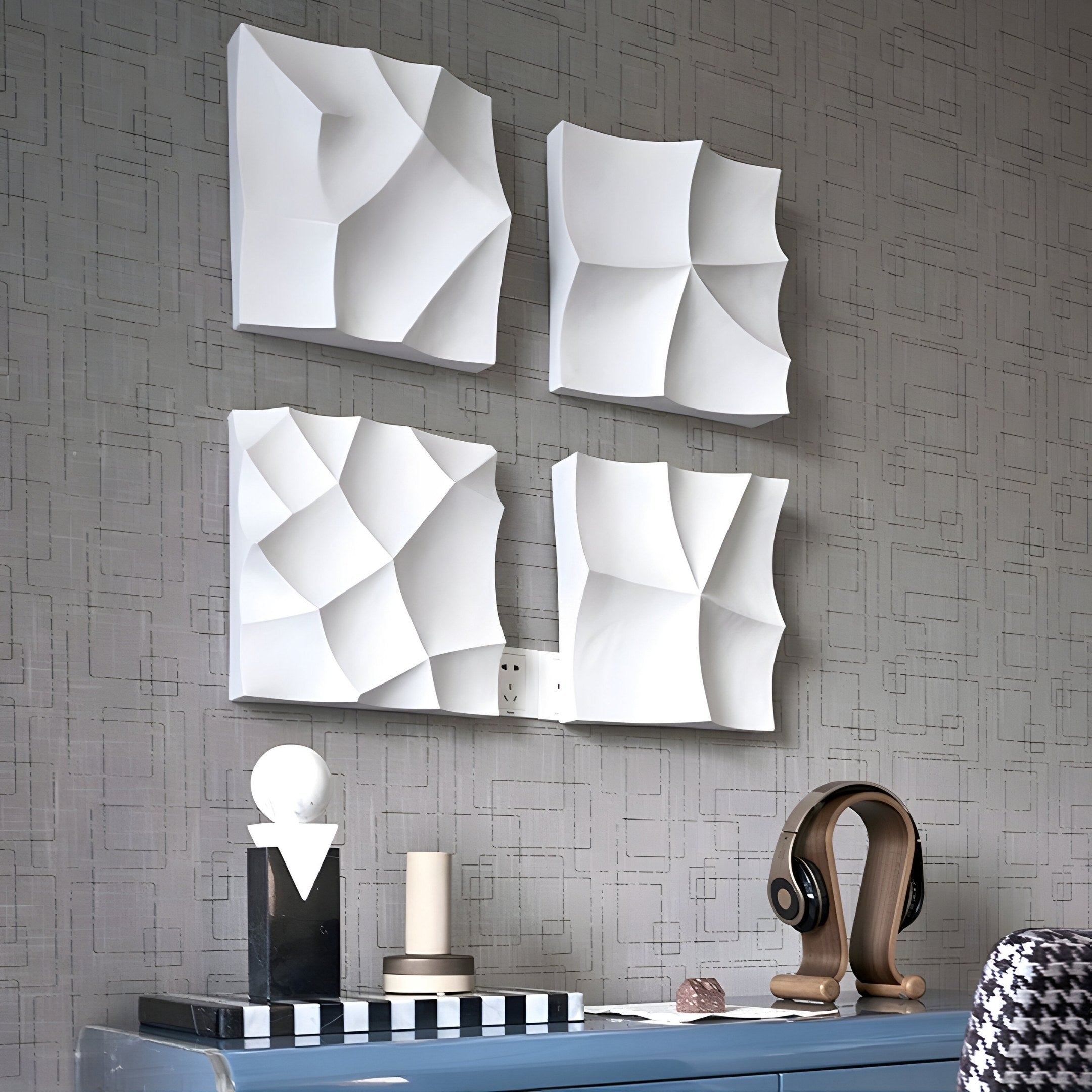 A modern interior highlights a textured gray wall with Giant Sculptures Tessara 4-Panel Set White 3D Wood Relief Wall Art, featuring abstract geometric panels. Below, a blue surface displays decorative objects like a sculpture and chic headphones on a stand, adding to the contemporary vibe.