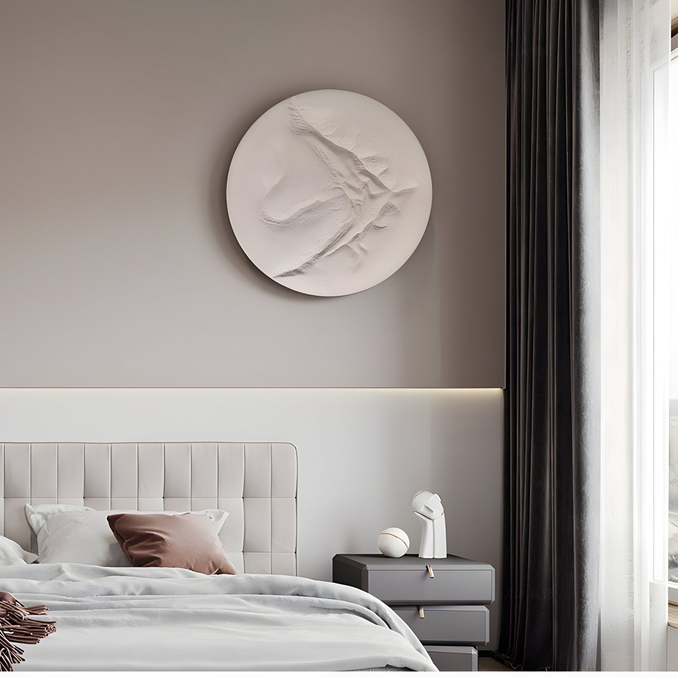 In a modern, minimalist bedroom, light gray bedding with a brown pillow complements the Selene I White Sandstone Textured 3D Wood Relief Wall Art by Giant Sculptures above. A dark curtain partially veils a large window beside a gray nightstand topped with a small sculpture and sphere.