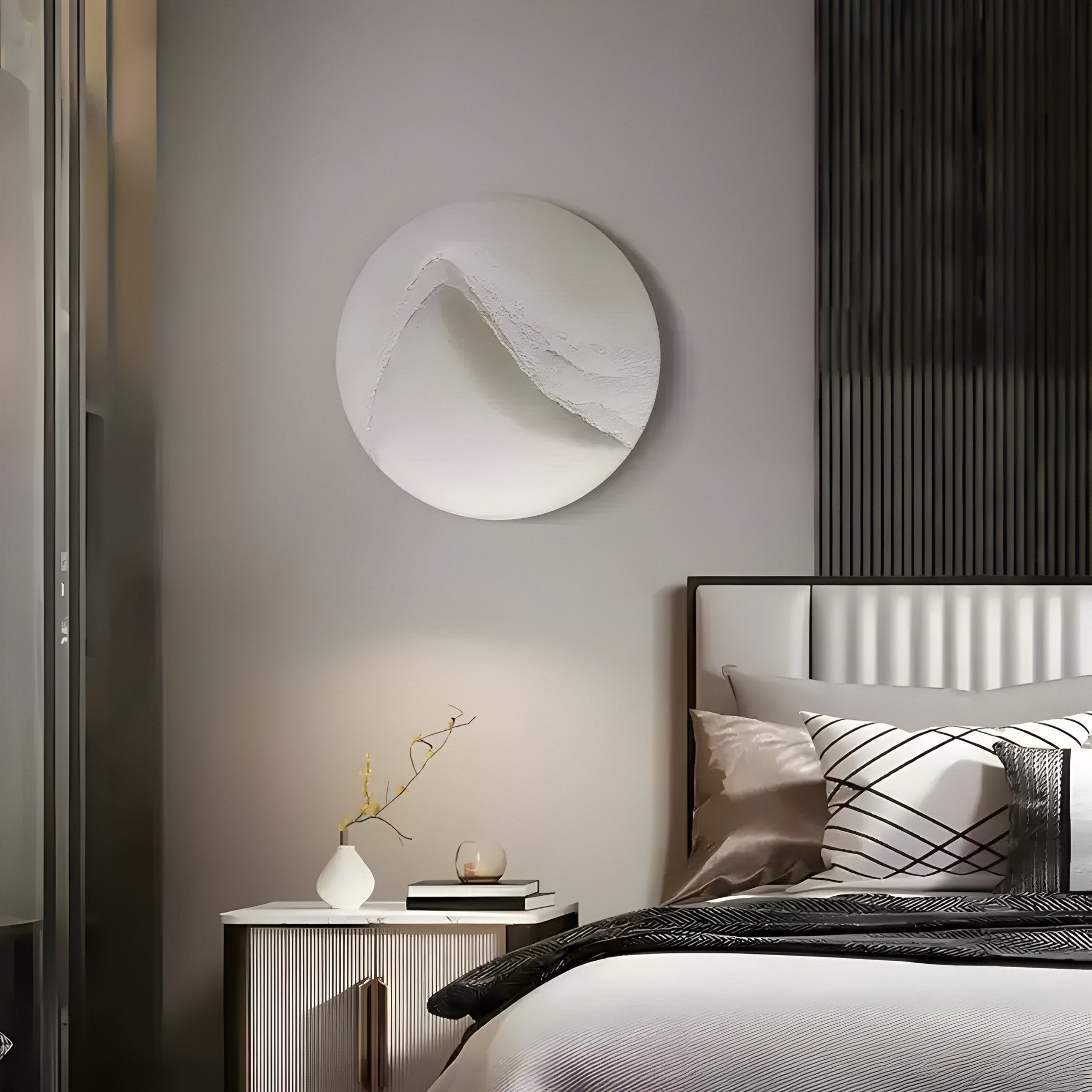 A modern bedroom features a tidy bed adorned with white and gray pillows and a dark throw blanket. Above the nightstand with a vase and candle, the Selene II White Sandstone Textured 3D Wood Relief Wall Art by Giant Sculptures enhances the light gray wall, adding depth to this minimalist sanctuary.
