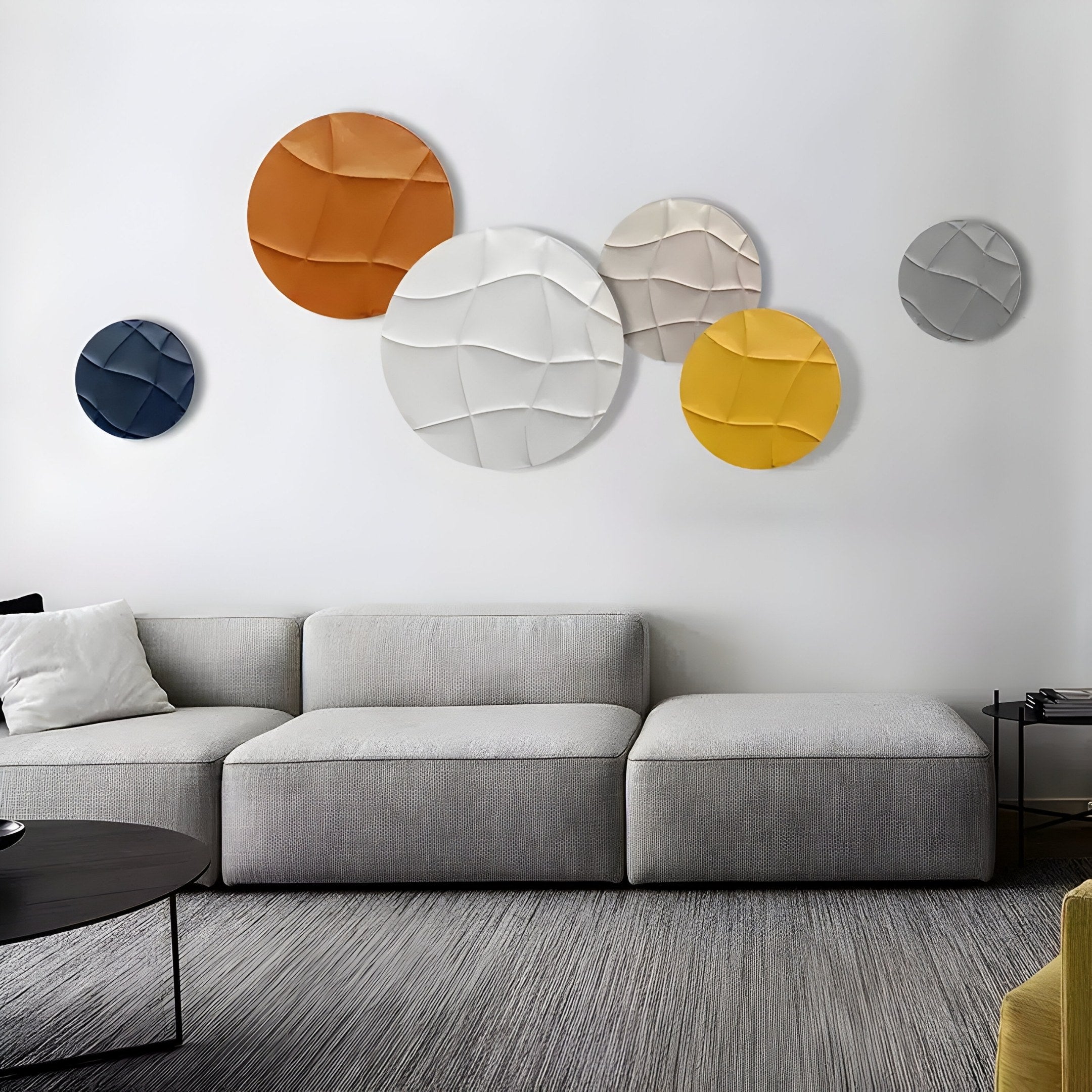 A modern living room showcases a gray sofa with white and dark cushions, a round black coffee table, and Giant Sculptures Orbita 6-Set Circular Earth-Tone 3D Wood Relief Wall Art in orange, white, yellow, and blue hues. The textured gray rug enhances the spaces coziness.