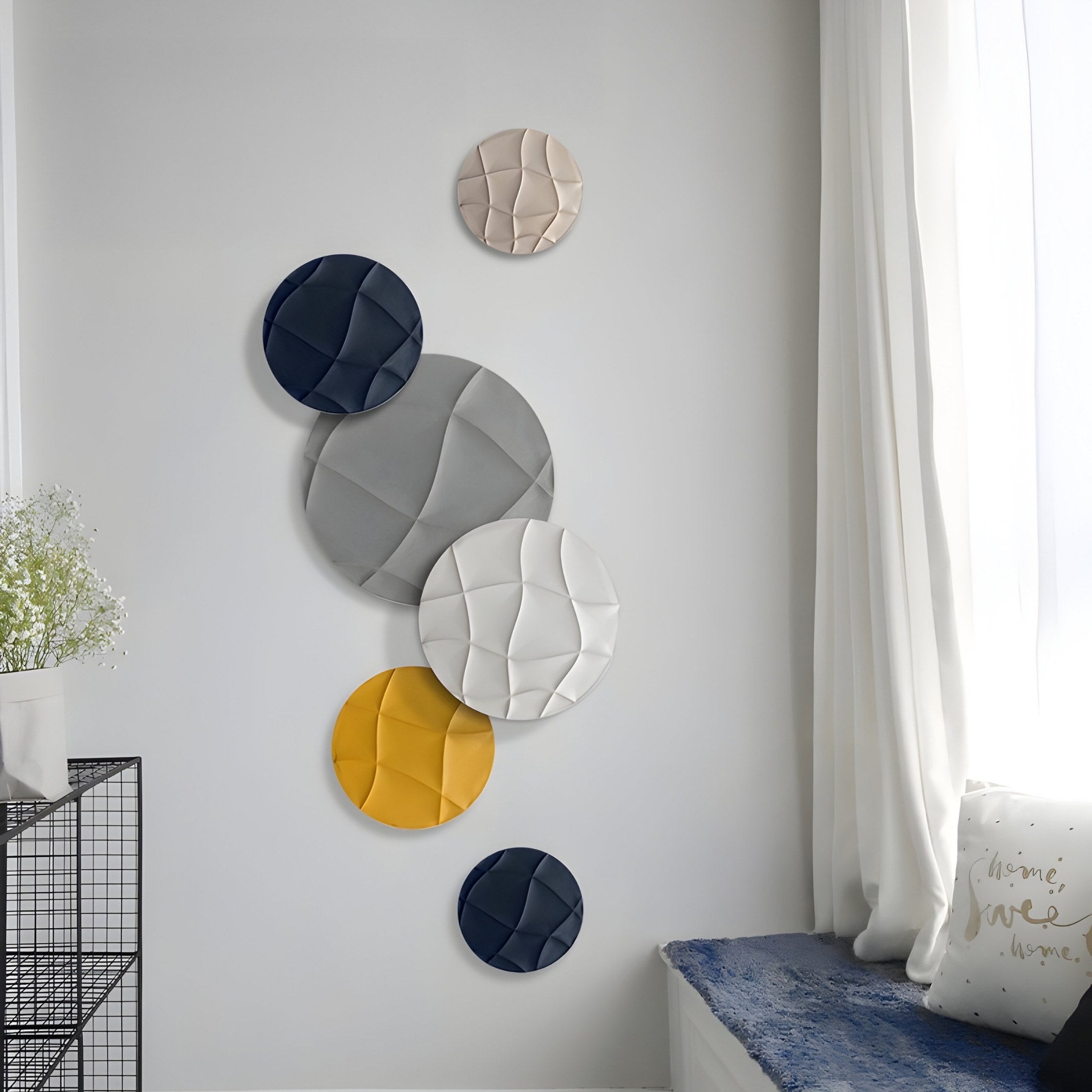 The Orbita 6-Set Circular Neutral Grey Wall Art by Giant Sculptures displays textured panels in black, white, beige, and yellow. With a sheer-curtained window nearby and a blue-cushioned bench plus wire-frame shelf with flowers, it enhances this stylish interior.