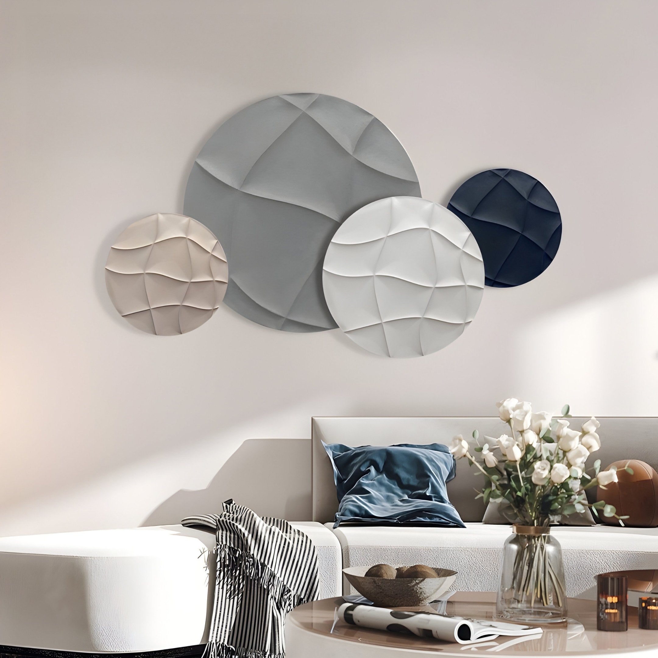 A modern living room features a sculptural white couch with blue and white cushions, a round white ottoman, and a vase of white flowers. The wall showcases the Orbita 4-Set Circular Monochrome 3D Wood Relief Wall Art by Giant Sculptures in shades of blue, gray, and beige.