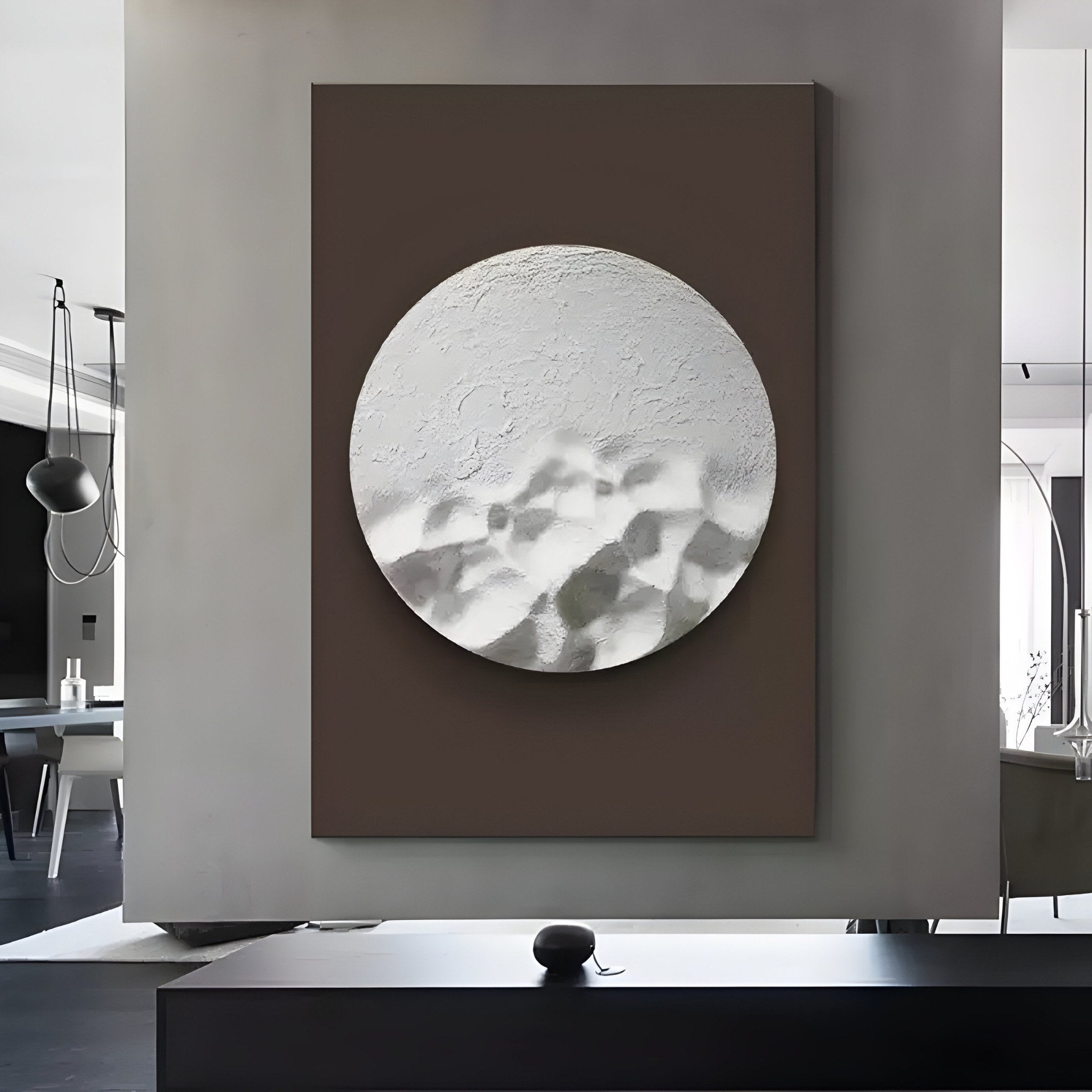 In a modern interior, the Solivis Arctic 3D Wood Relief Wall Art by Giant Sculptures displays a textured white circular design on brown, enhancing a sleek monochrome space with contemporary furnishings and pendant lights.