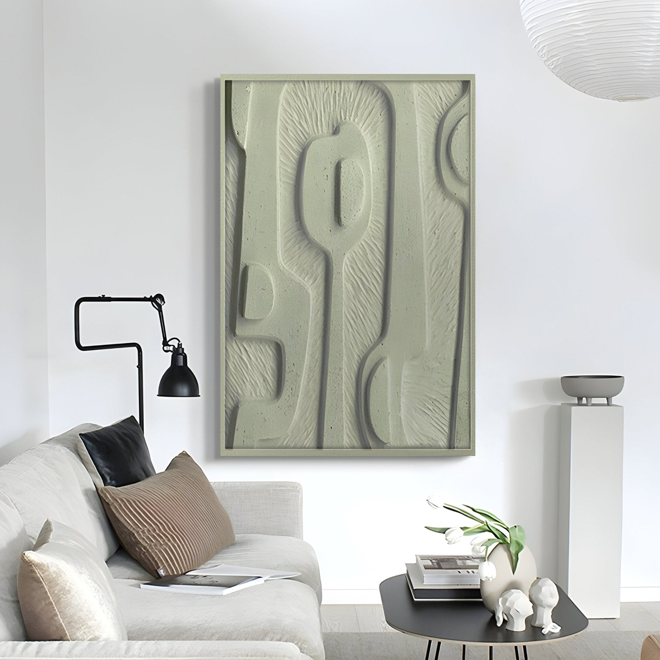 In a modern living room, a white sofa with black and brown cushions sits beneath the Terravura Rectangular Sage Green I 3D Wood Relief Wall Art by Giant Sculptures. Beside it, a sleek black lamp stands tall, while a small table with books and lush greenery enhances the natural decor.