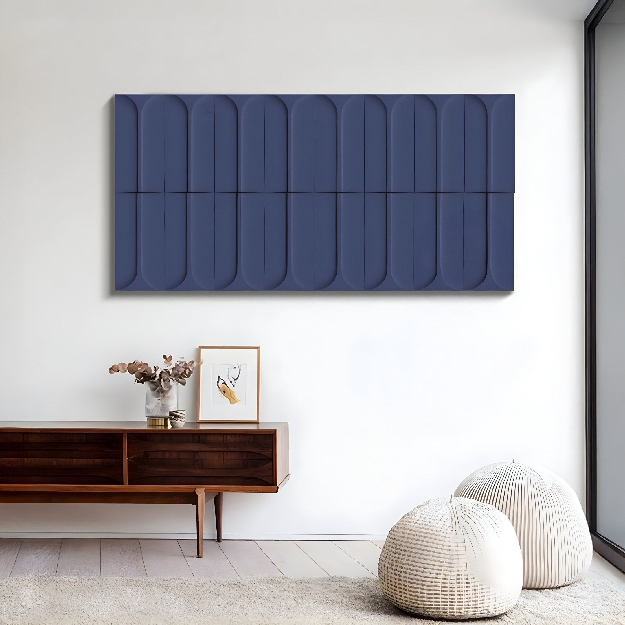 A minimalist room includes a wooden console table, a framed abstract line drawing, and a vase with dried flowers. Two white poufs are on the floor. The Vireus Midnight Blue Rectangular 3D Wood Relief Wall Panel by Giant Sculptures hangs above, while natural light floods in through a large window.